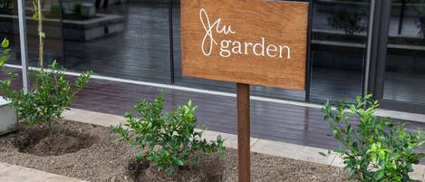 A wooden sign with the text "Ju garden" in front of a building with glass windows and a paved area with small plants.