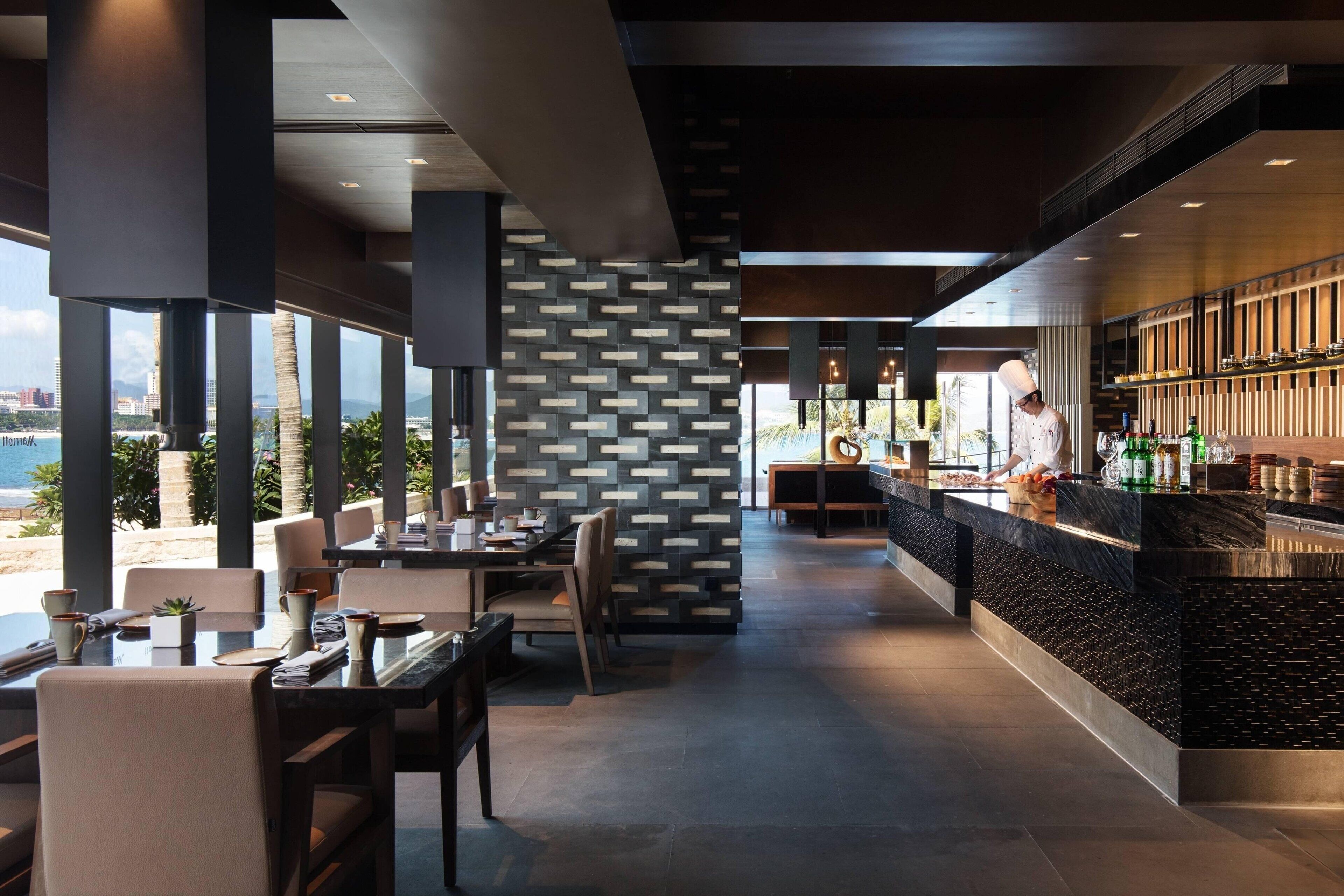 Een modern restaurant met een kok die gerechten bereidt, grote ramen met uitzicht op een strand en een stijlvol interieur.