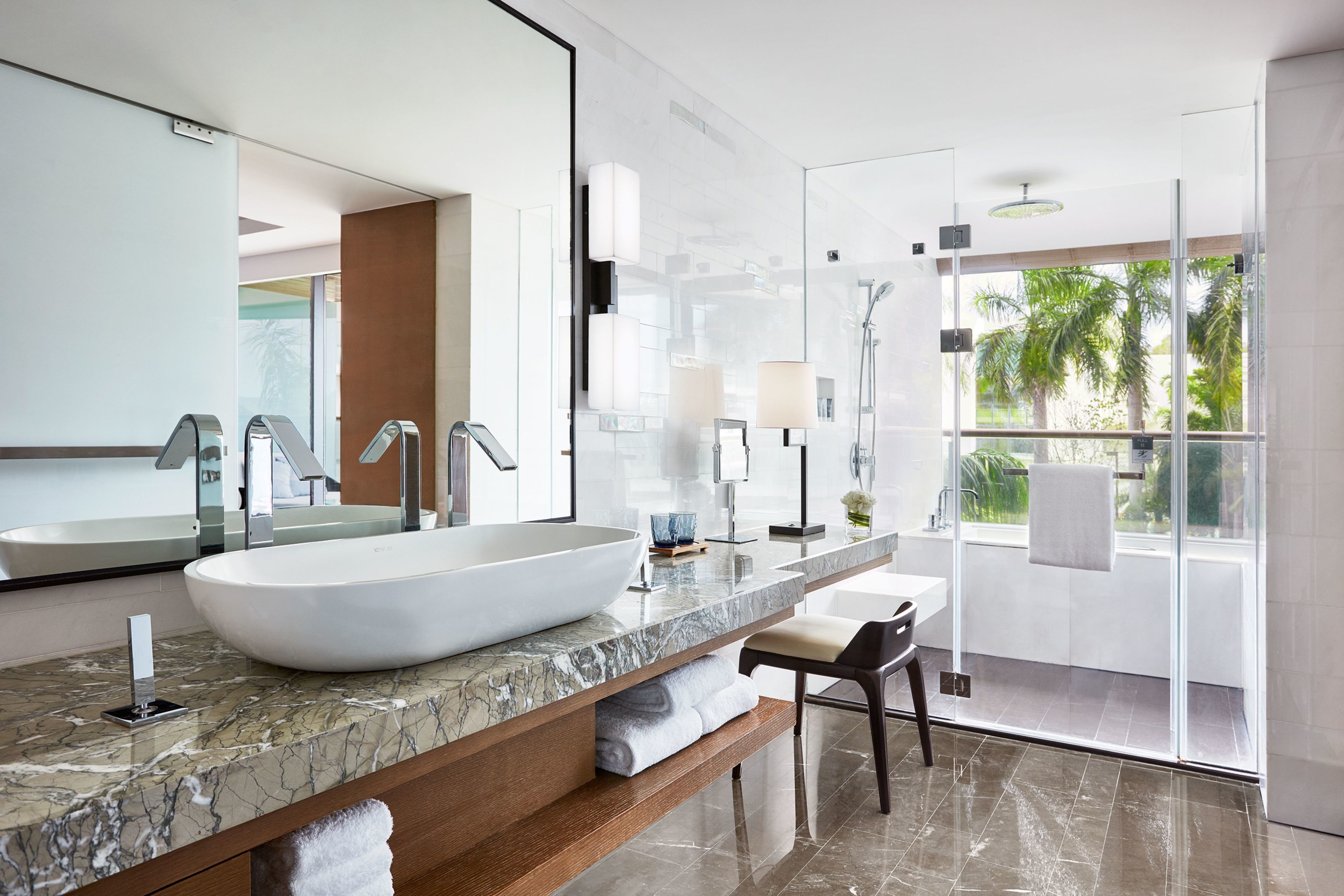 A modern bathroom with a large white sink, a marble countertop, a wooden vanity, a large mirror, and a glass shower enclosure with a view of palm trees.