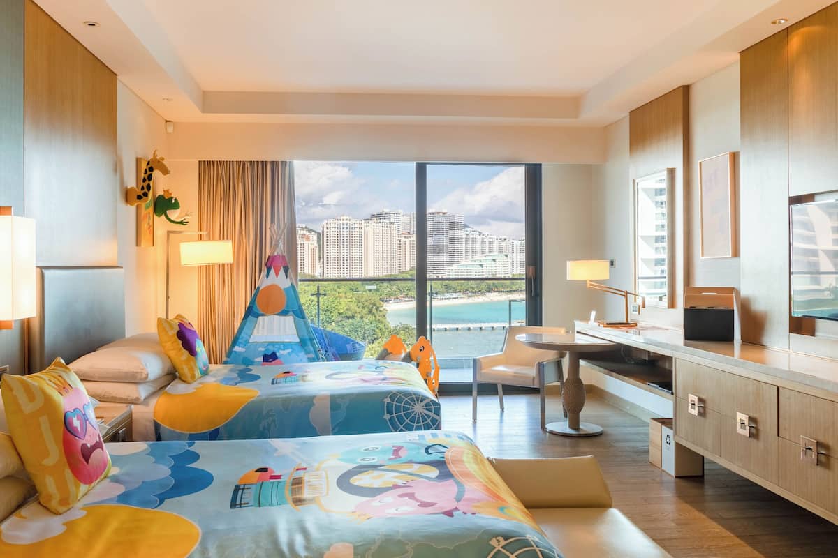 A hotel room with two beds, a desk, a chair, and a large window with a city view.