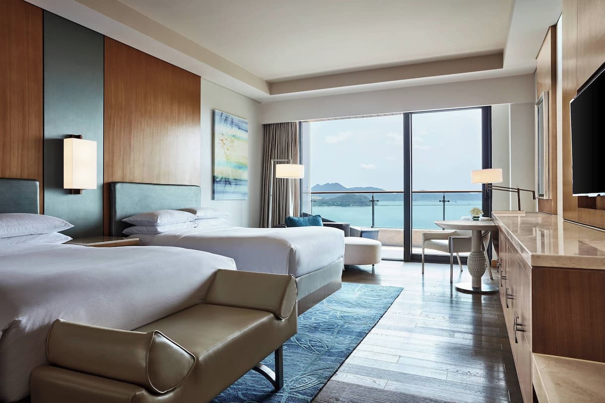 A hotel room with two beds, a sofa, a dining area with a table and chairs, a TV, and a large window with a view of the sea.