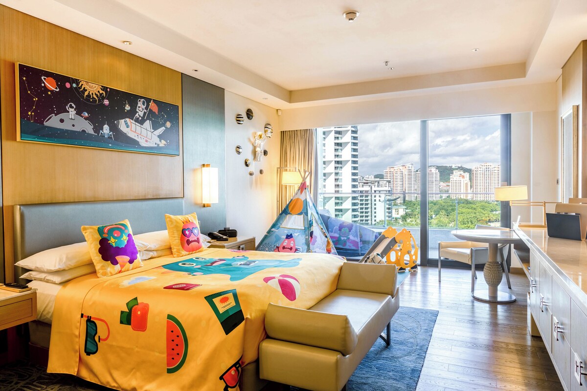 A hotel room with a large bed, a desk, a chair, a window with a city view, and a painting on the wall.