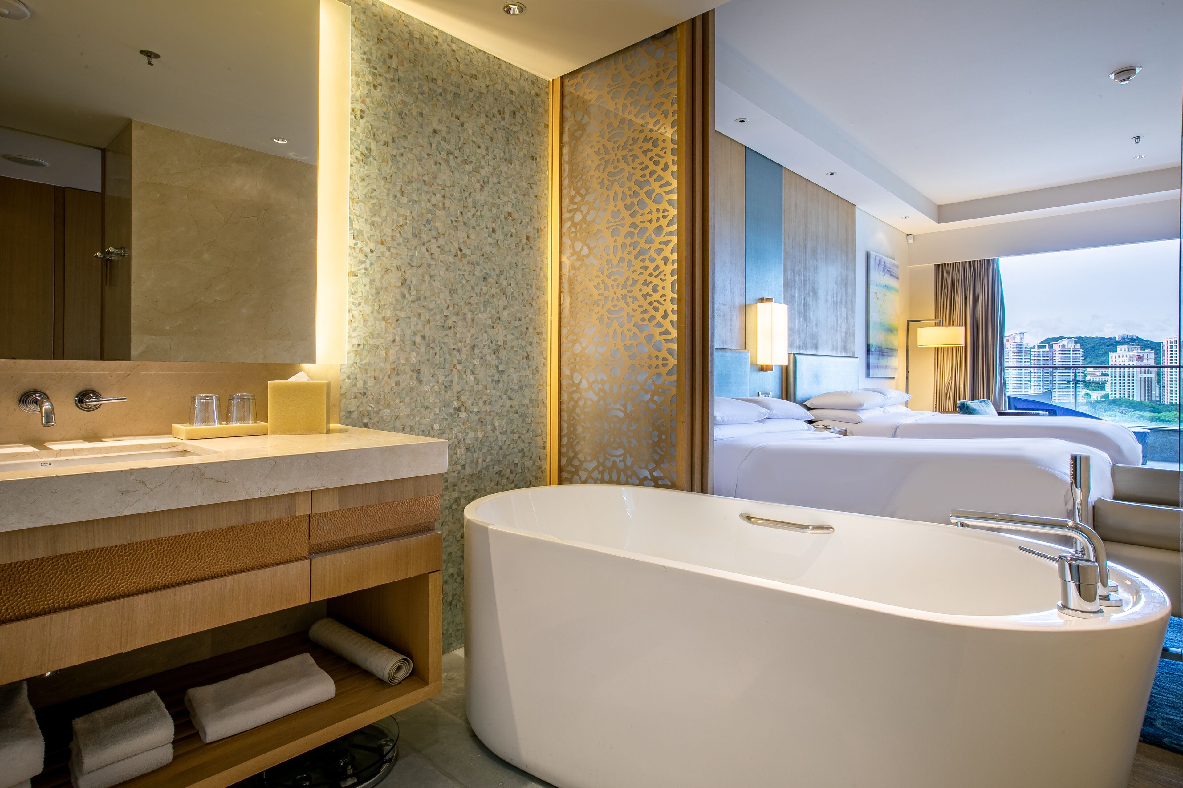 A modern hotel bathroom with a large bathtub, a vanity with a mirror, and a view of the city through large windows.