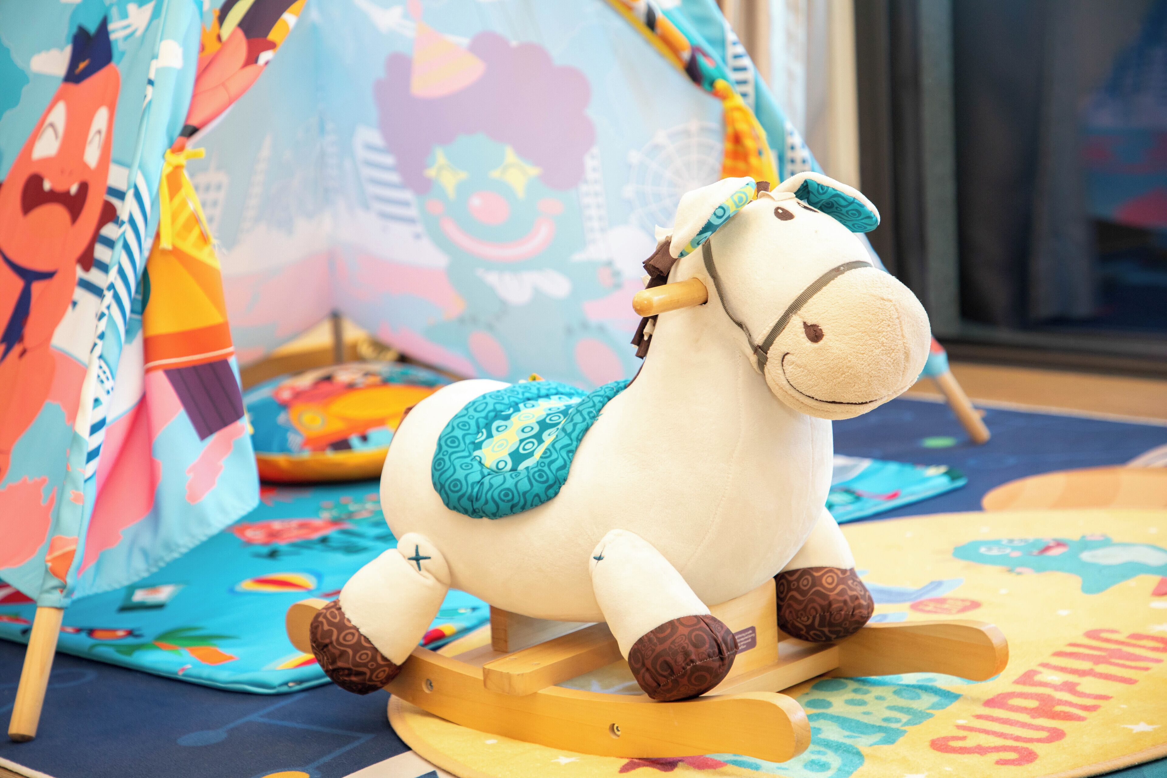 A white rocking horse with brown accents and a blue blanket, placed on a colorful mat with a 'Surfing' theme.