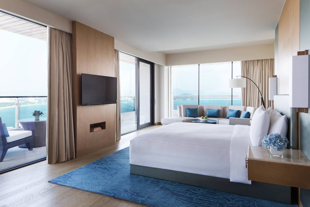 A modern hotel room with a large bed, a TV mounted on the wall, a balcony with a view of the sea, and a seating area with a sofa and chairs.