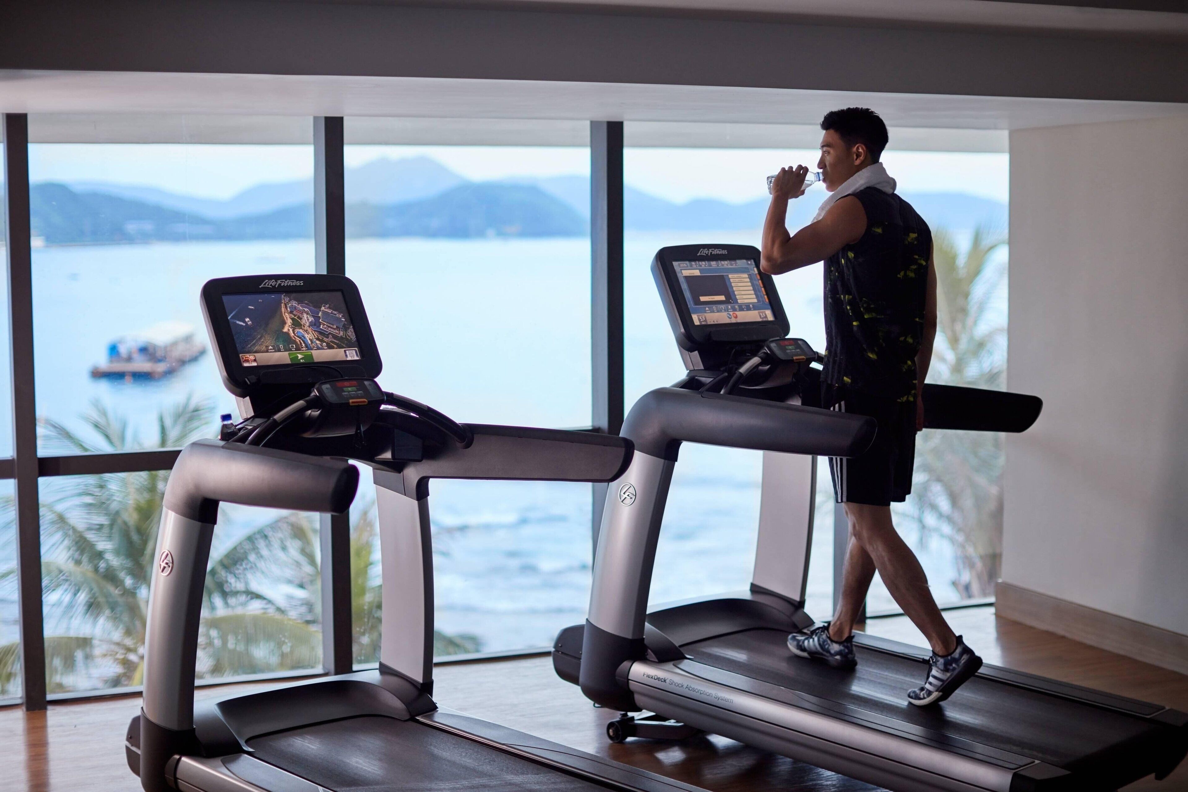 A person using a treadmill with a digital display, in a room with large windows overlooking a body of water and palm trees.