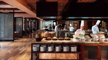A modern kitchen with chefs preparing food, wooden flooring, and a buffet area with various dishes.