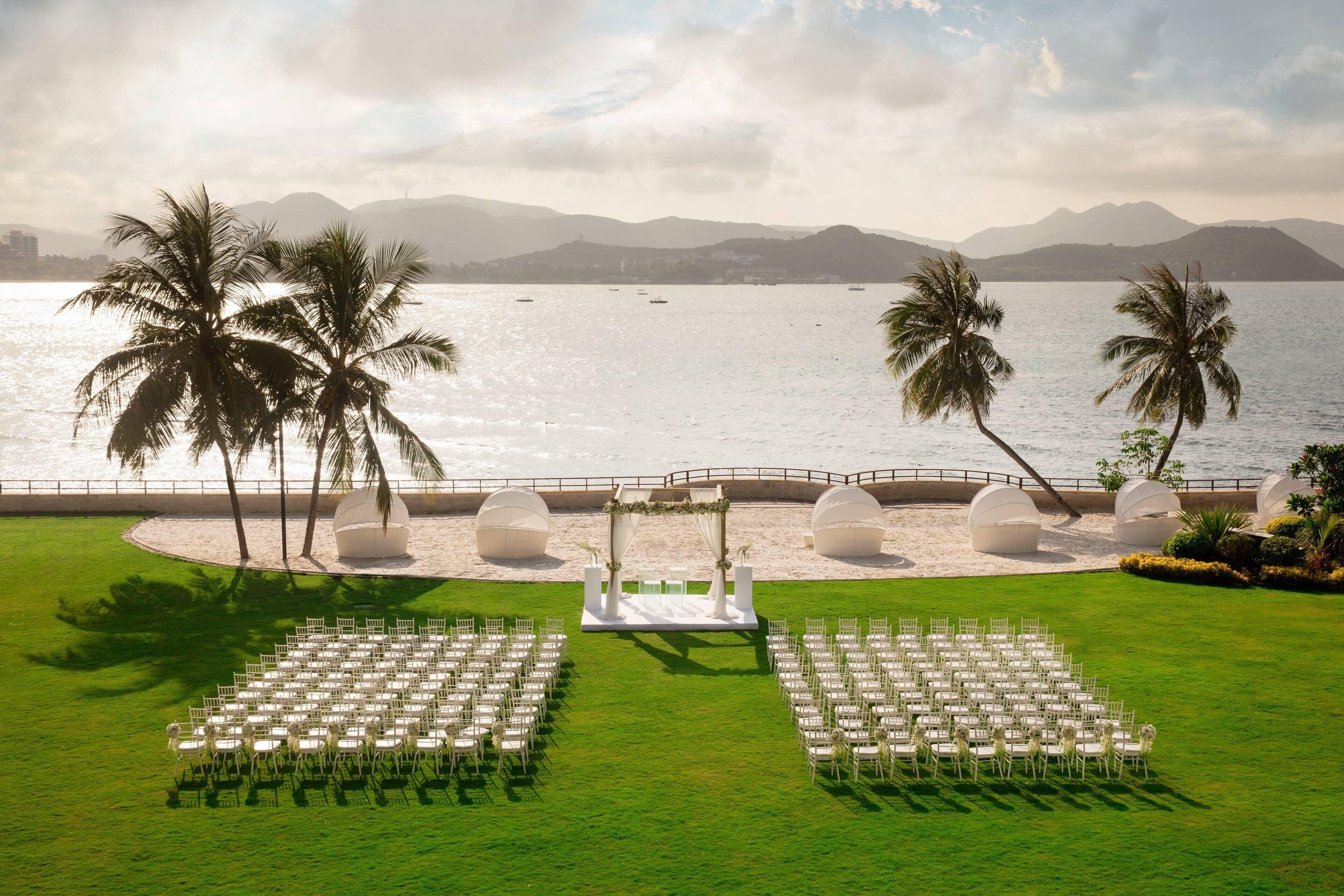An outdoor wedding ceremony setup with a stage, chairs, and palm trees by a beach.