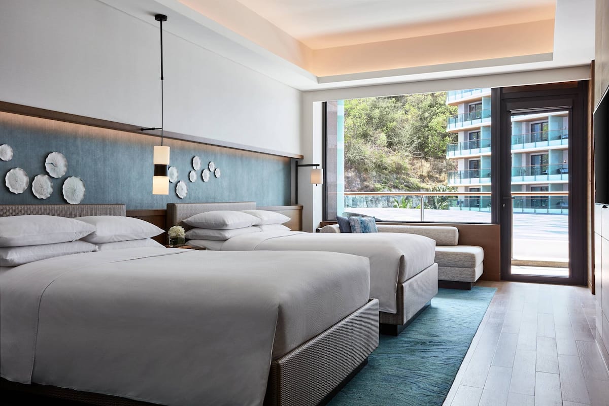 A modern hotel room with two beds, a large window with a view of a building, and a balcony.