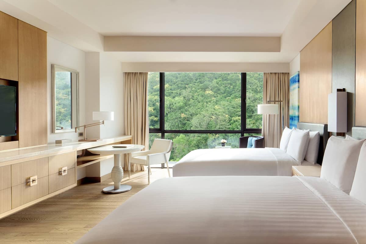 A hotel room with two beds, a desk, a chair, a TV, and a large window with a forest view.