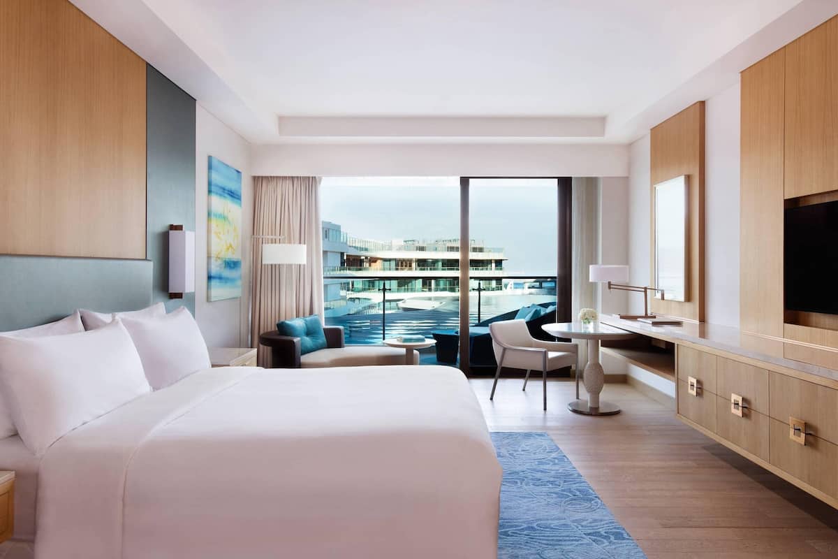 A hotel room with a large bed, a desk, a chair, and a view of a building and water.