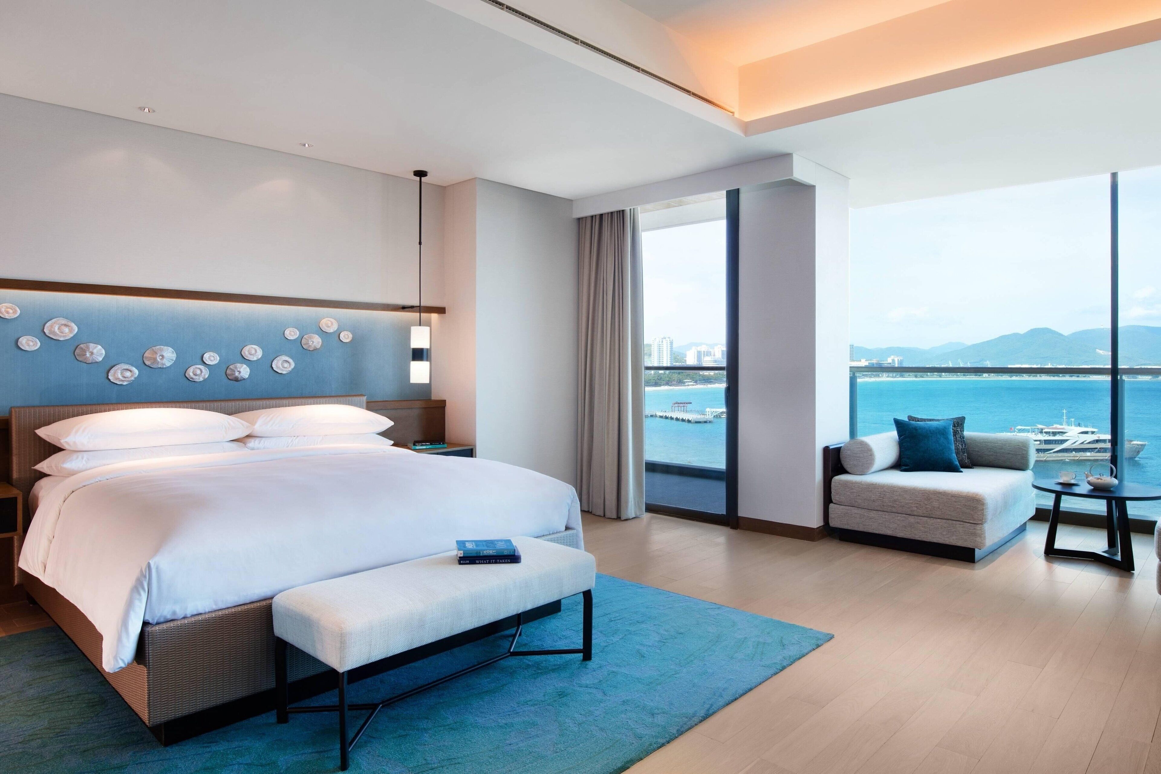 A modern hotel room with a large bed, a sofa, a small table, and a view of the sea.