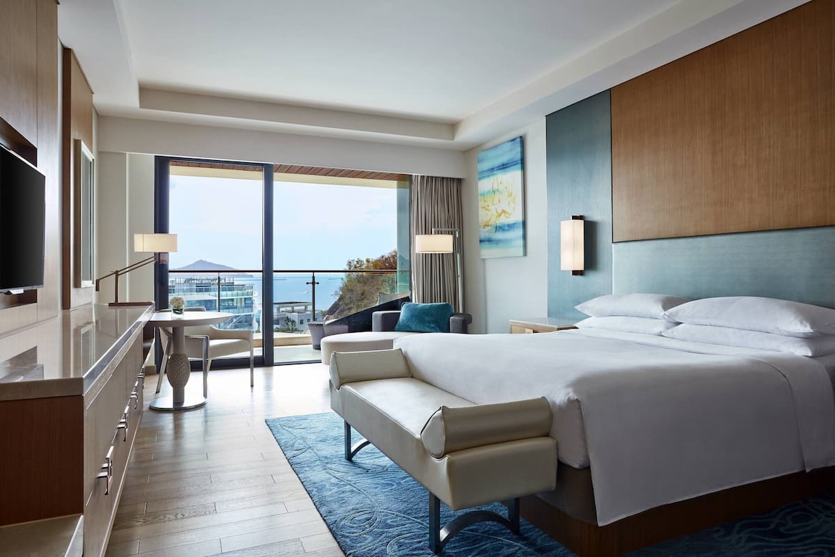 A modern hotel room with a large bed, a sofa, a dining area, and a balcony with a view.