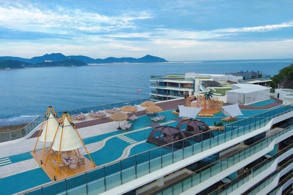 A cruise ship deck with a swimming pool, sun loungers, and a view of the ocean and distant mountains.