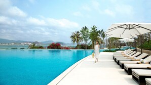 Zona junto a la piscina con sillones de descanso y sombrillas, cielo despejado y vista al mar y montañas al fondo.
