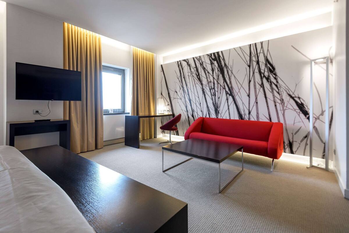 Ein modernes Hotelzimmer mit einem an der Wand befestigten Flachbildfernseher, einem roten Sofa, einem dunklen Couchtisch und einem großen Fenster mit Vorhängen.