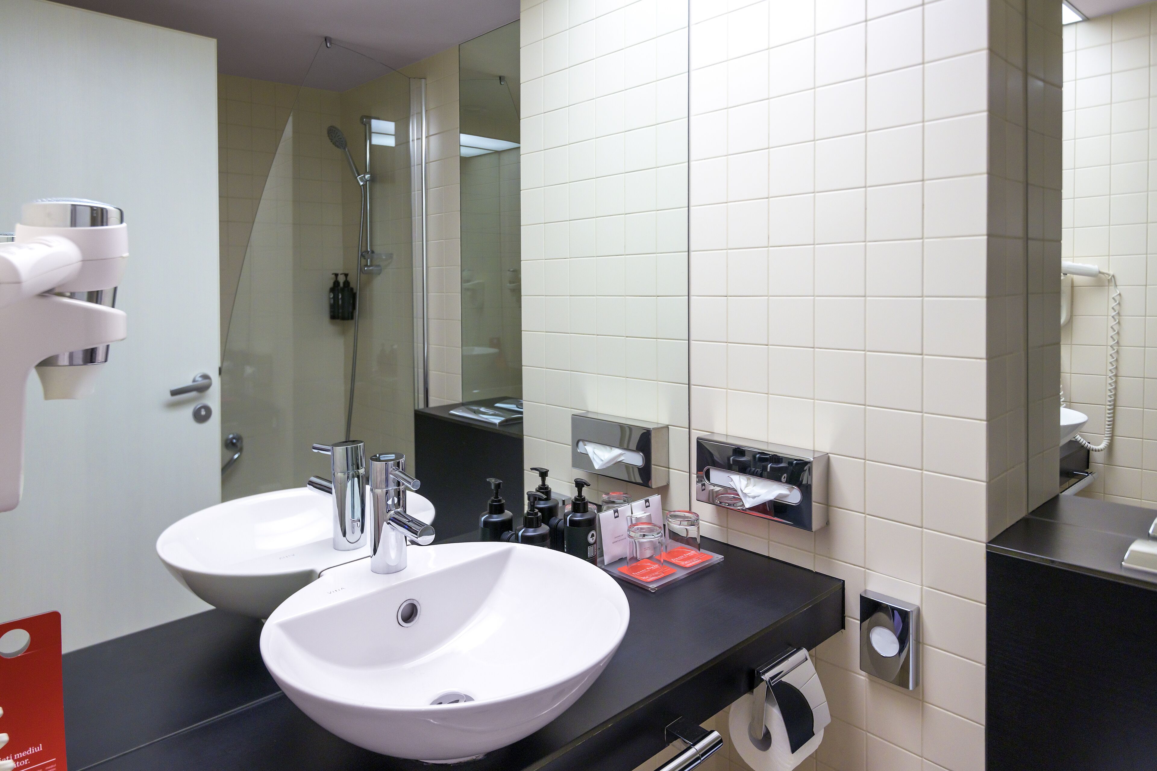 A modern bathroom with two sinks, a glass shower enclosure, and a towel dispenser.