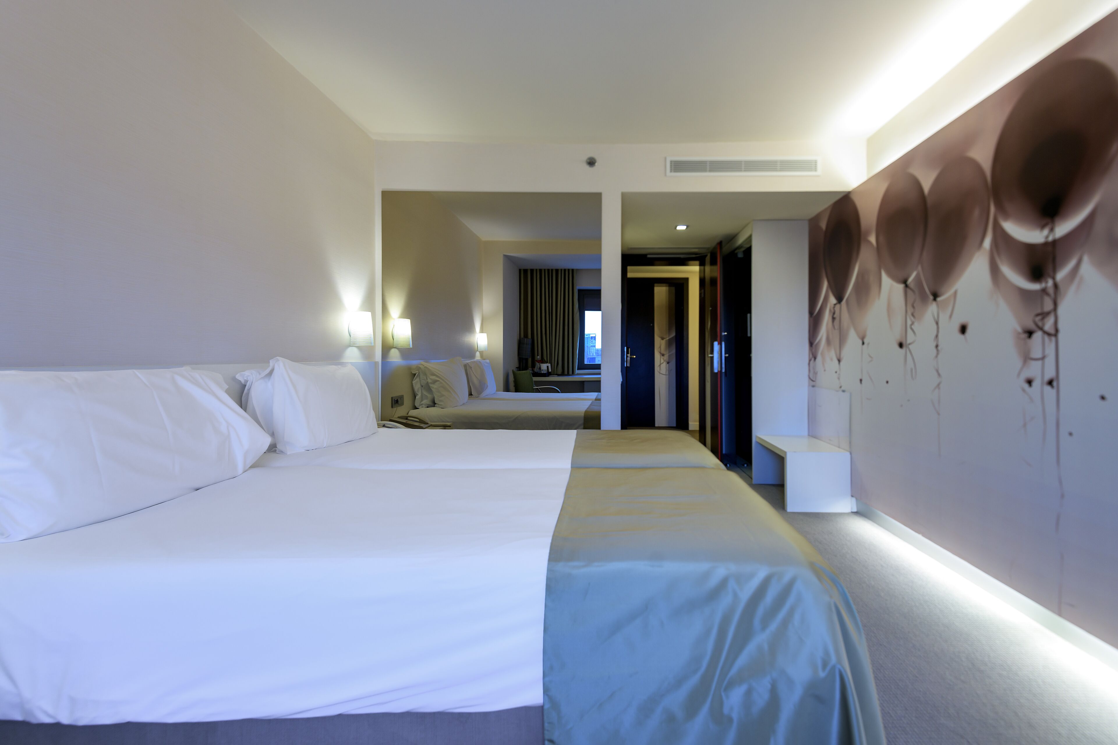 Habitación de hotel con dos camas, una pared grande con un mural de globos y un pasillo que conduce a otra habitación.