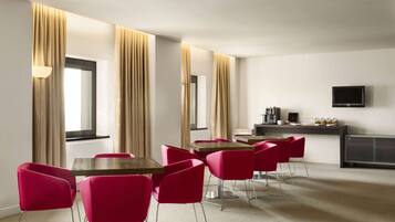A hotel room with a dining area featuring red chairs and wooden tables, a flat-screen TV, and a minibar.