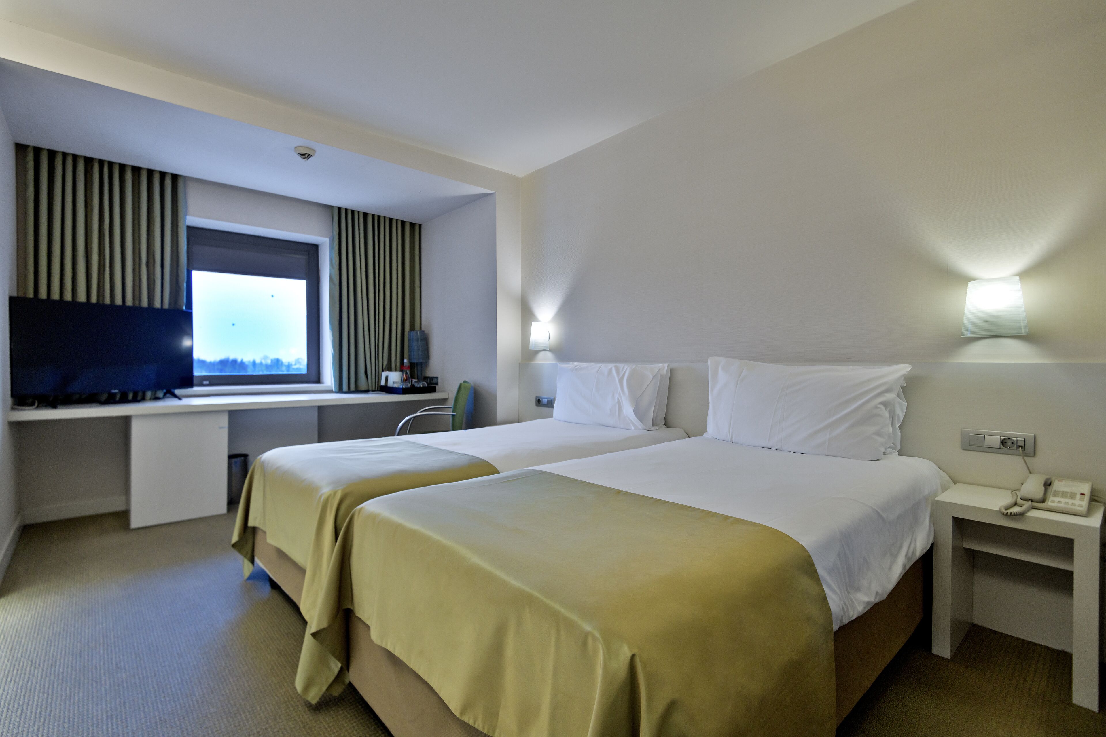 A hotel room with two beds, a desk, a TV, and a window with curtains.