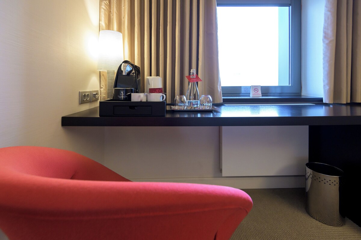 Una habitación de hotel con escritorio, lámpara, cafetera y ventana con cortinas.