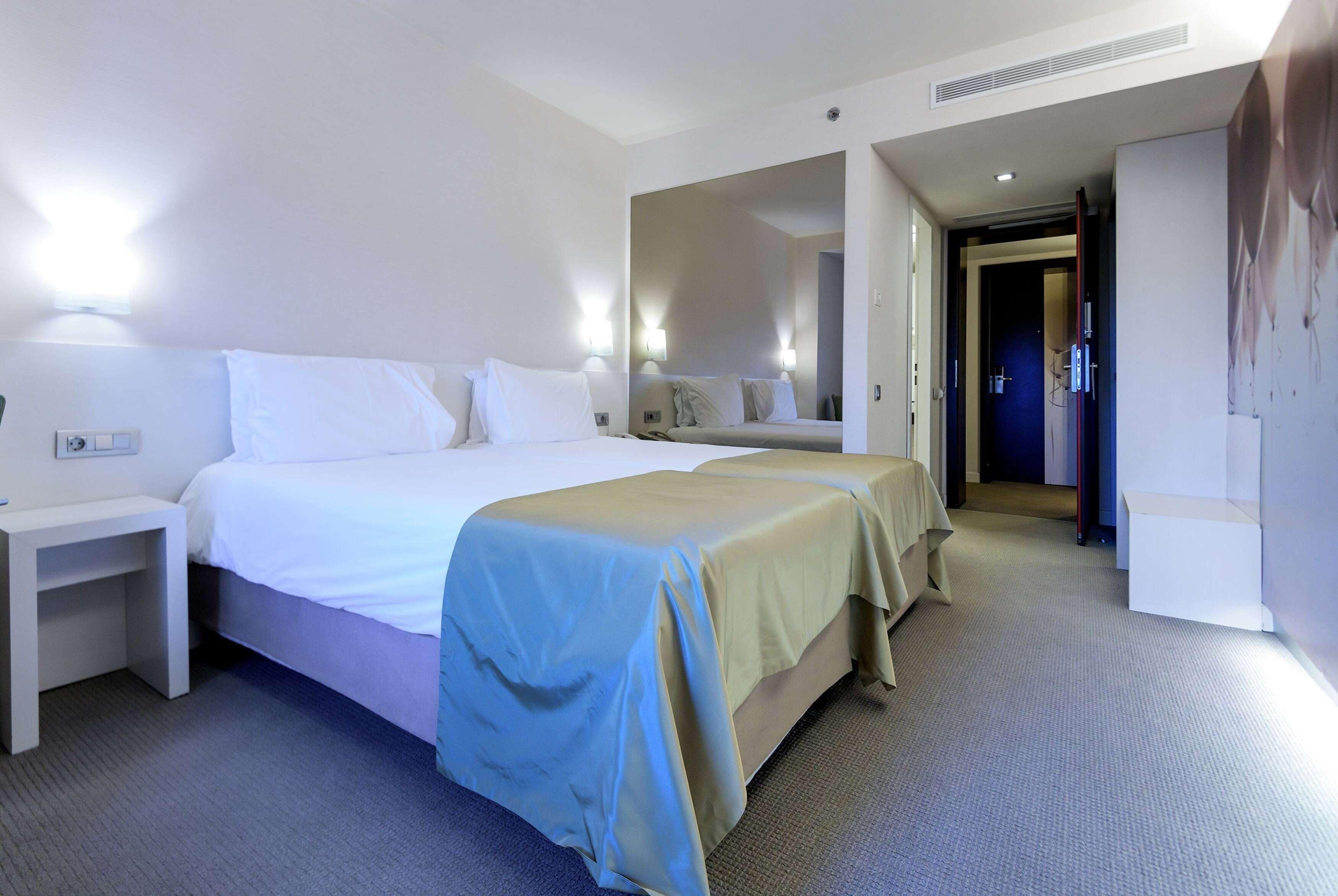 Habitación de hotel con dos camas, una mesita de noche, un espejo y una puerta que da a otra habitación.