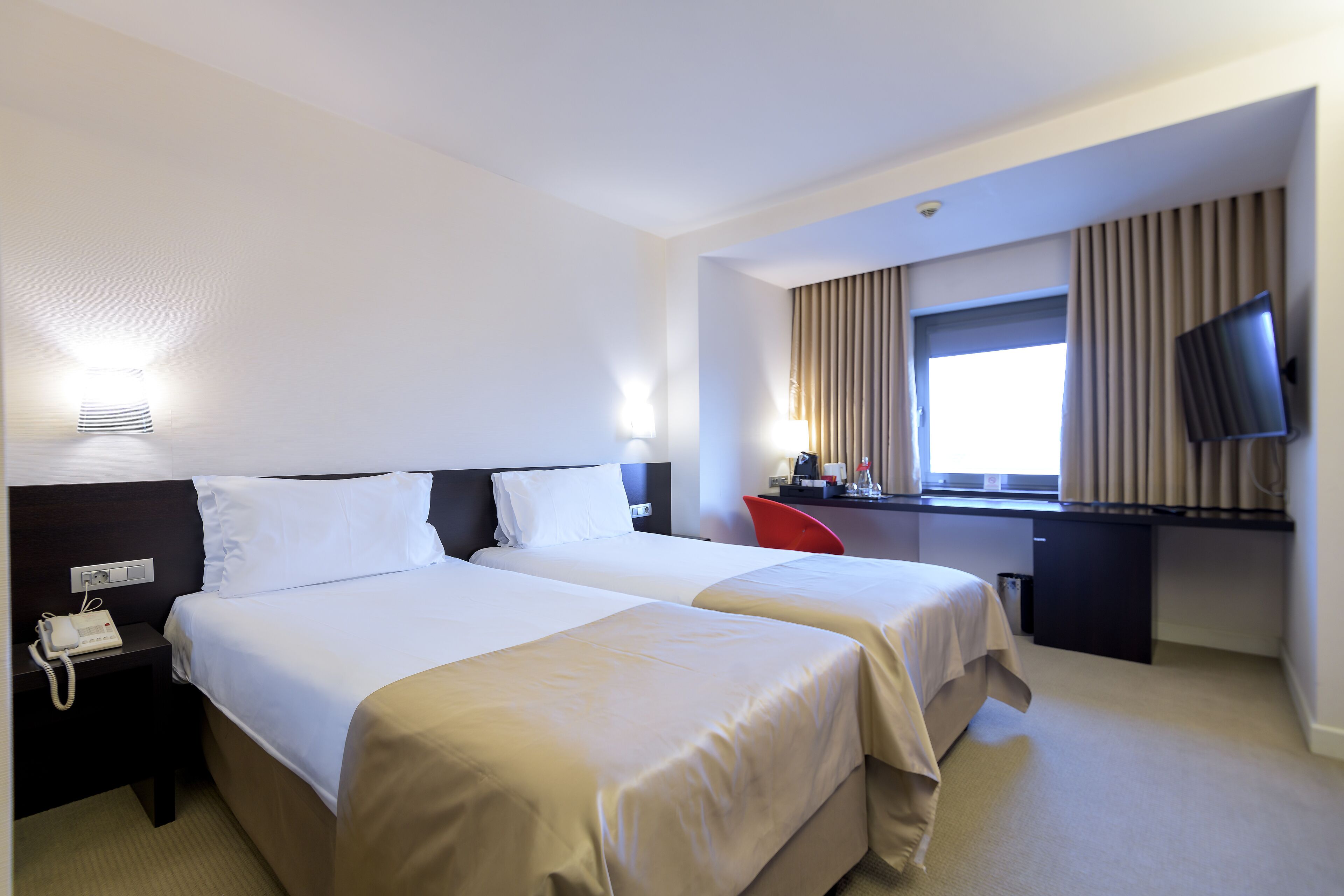 Quarto de hotel com duas camas, uma mesa, uma televisão e uma janela com cortinas.