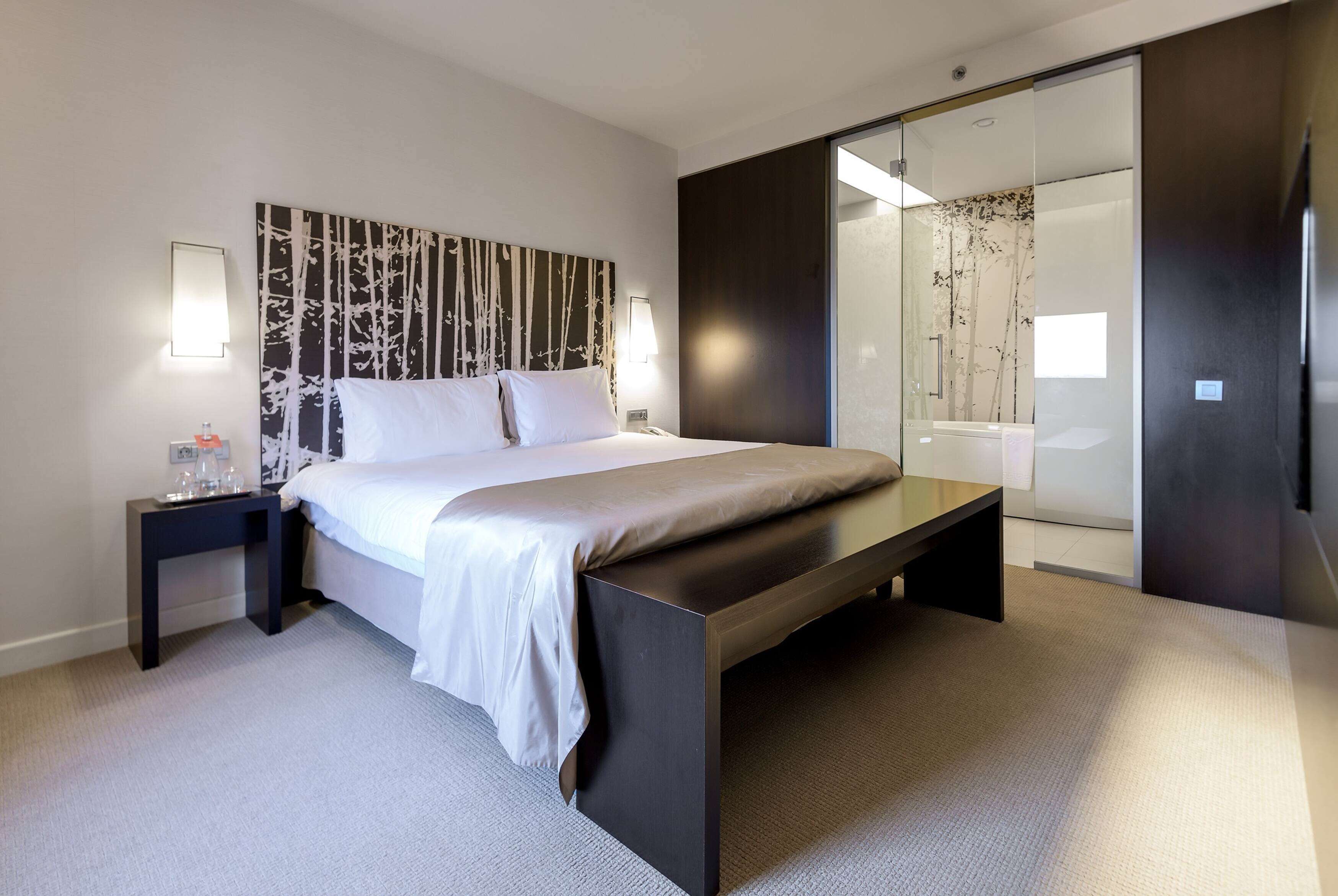 Una habitación de hotel moderna con una cama grande, una mesita de noche con lámpara, un box de ducha de vidrio y un banco de color oscuro.