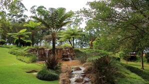A lush garden with a small waterfall, green grass, and various trees. - Greenway Woods Resort (Mbombela)
