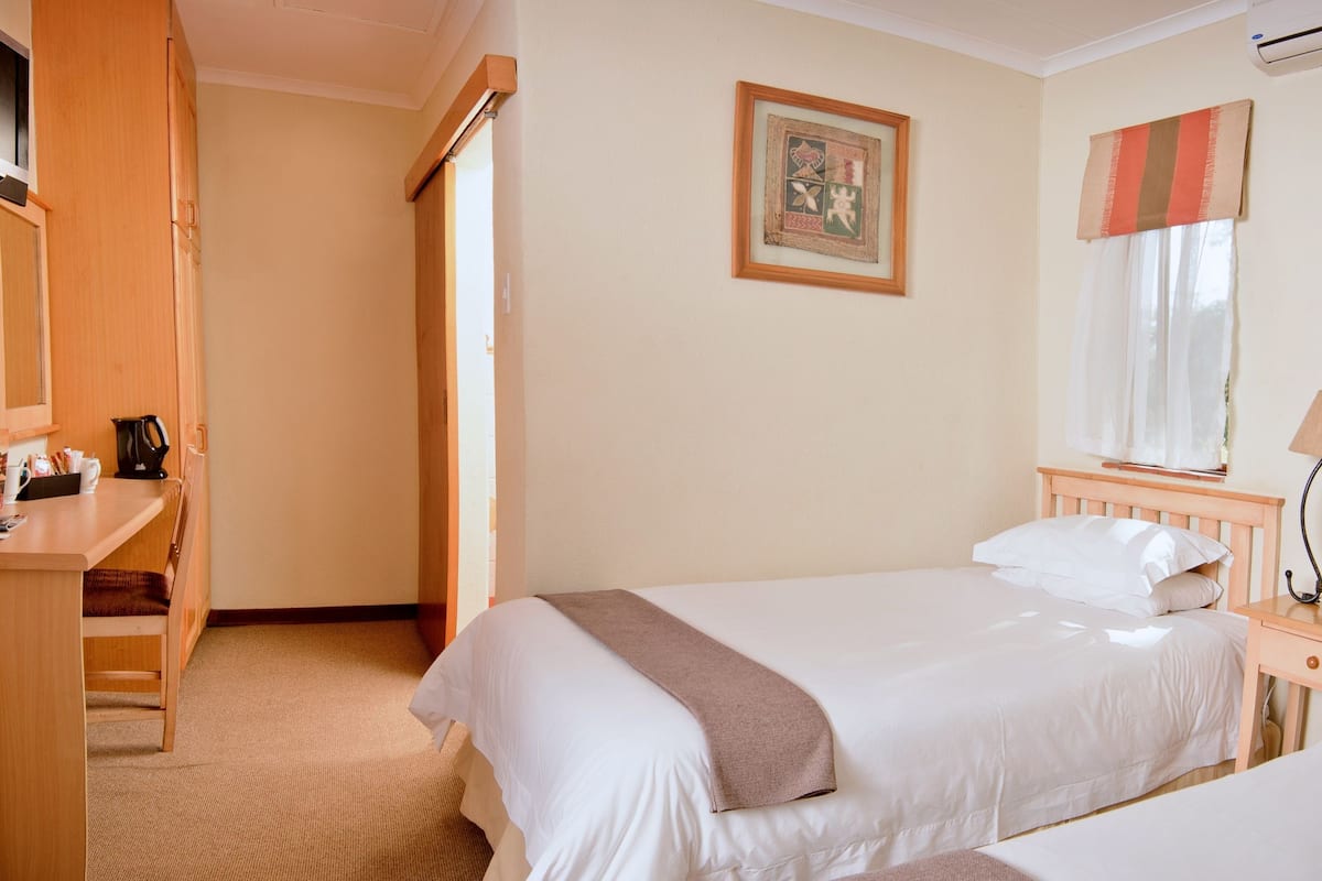 A hotel room with two beds, a desk with a chair, a bedside table with a lamp, and a framed picture on the wall.