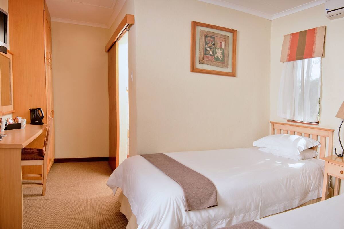 A hotel room with two beds, a desk, a chair, a bedside table with a lamp, and an air conditioning unit.