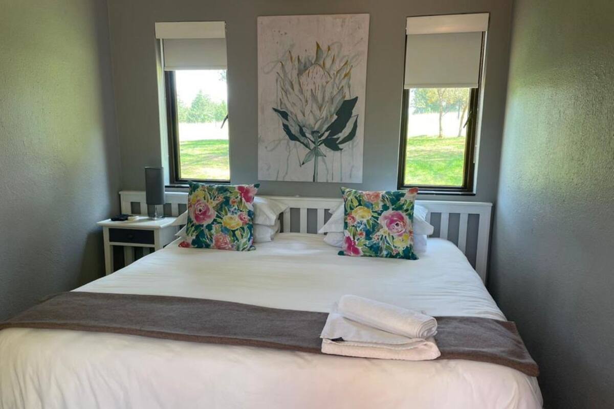 A neatly made bed with floral pillows, a white comforter, and a folded towel. There are two windows with blinds, a nightstand with a lamp, and a painting on the wall.