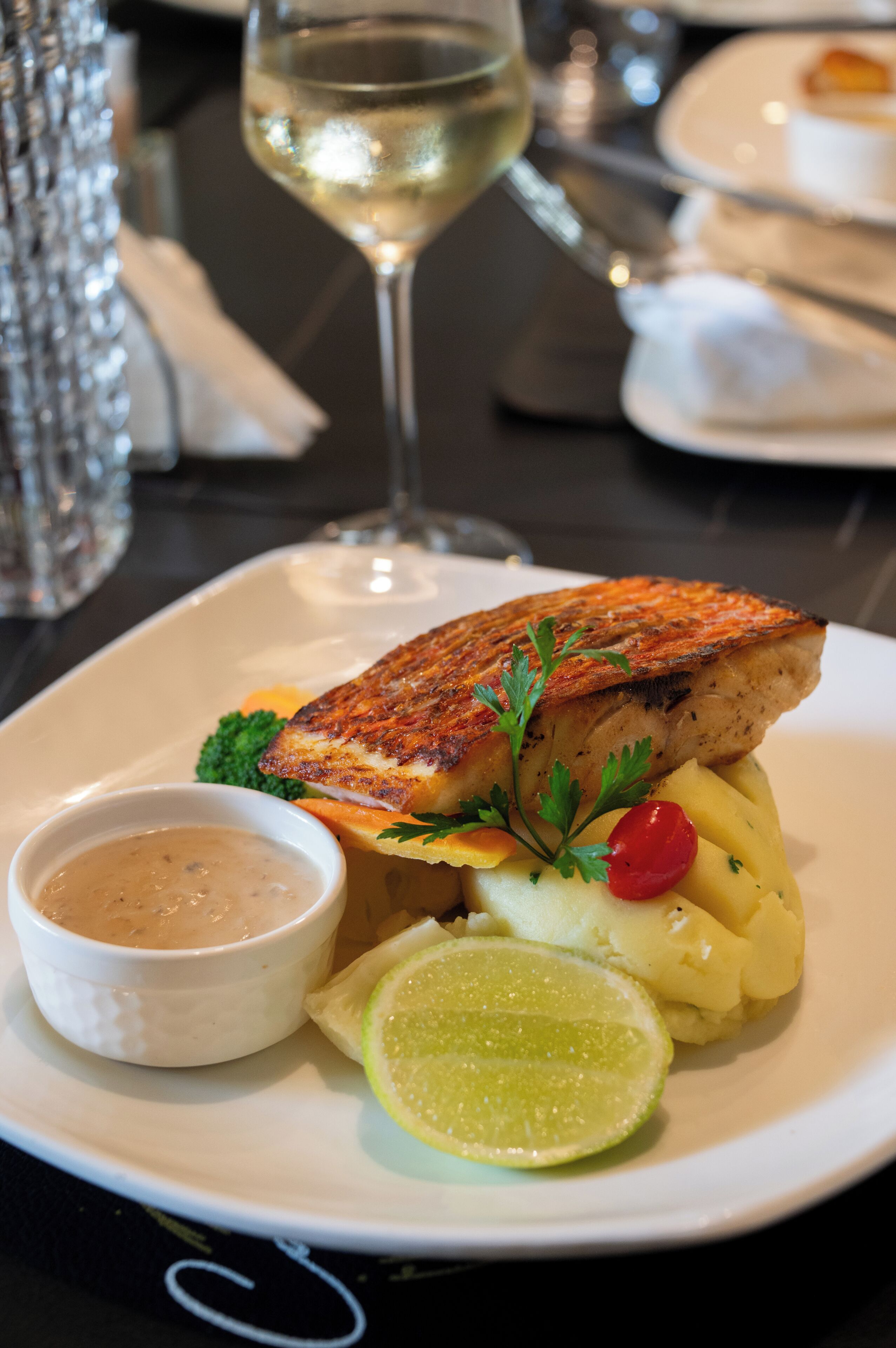 Un plato con pescado a la parrilla, puré de papas, una cuña de lima y un recipiente pequeño con salsa, junto a un vaso de vino blanco al fondo.