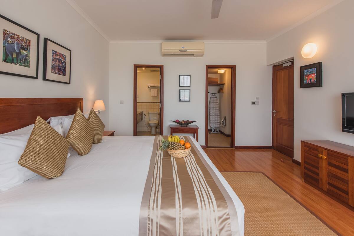 A hotel room with a large bed, a bedside table with a fruit bowl, a wooden headboard, and a TV on a wooden cabinet.