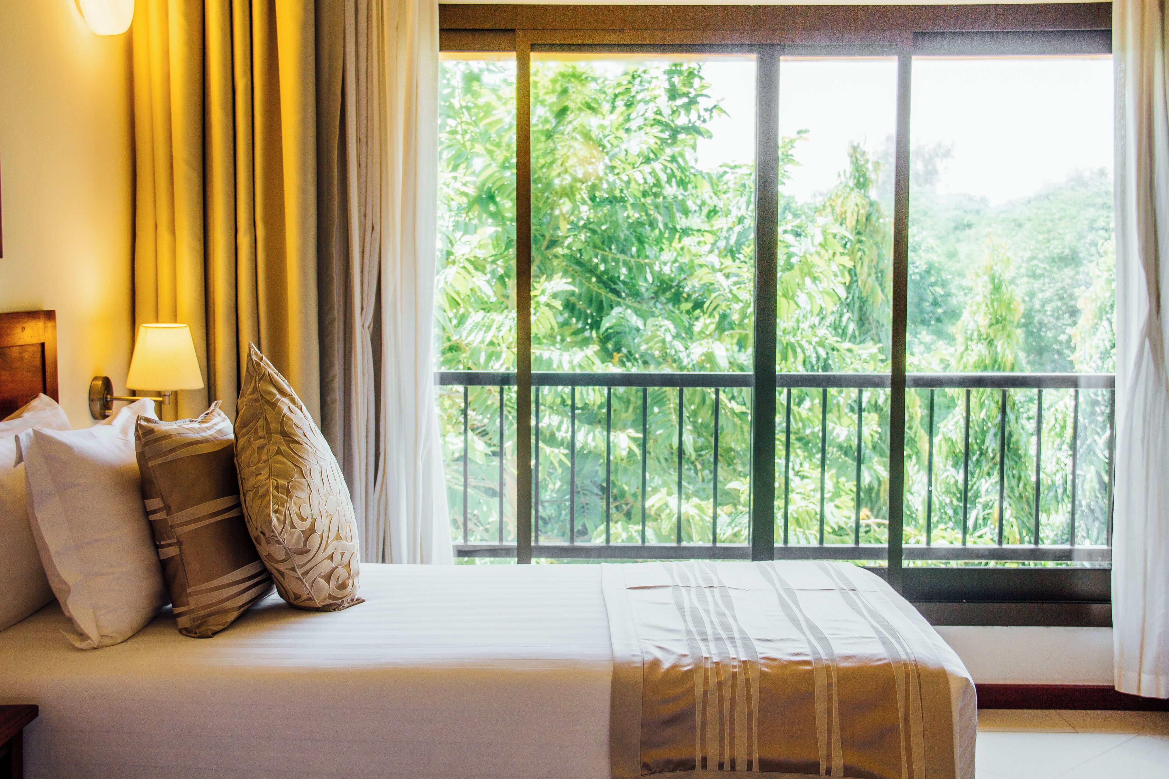 A hotel room with a bed, pillows, a nightstand, a lamp, and a large window with a view of greenery.