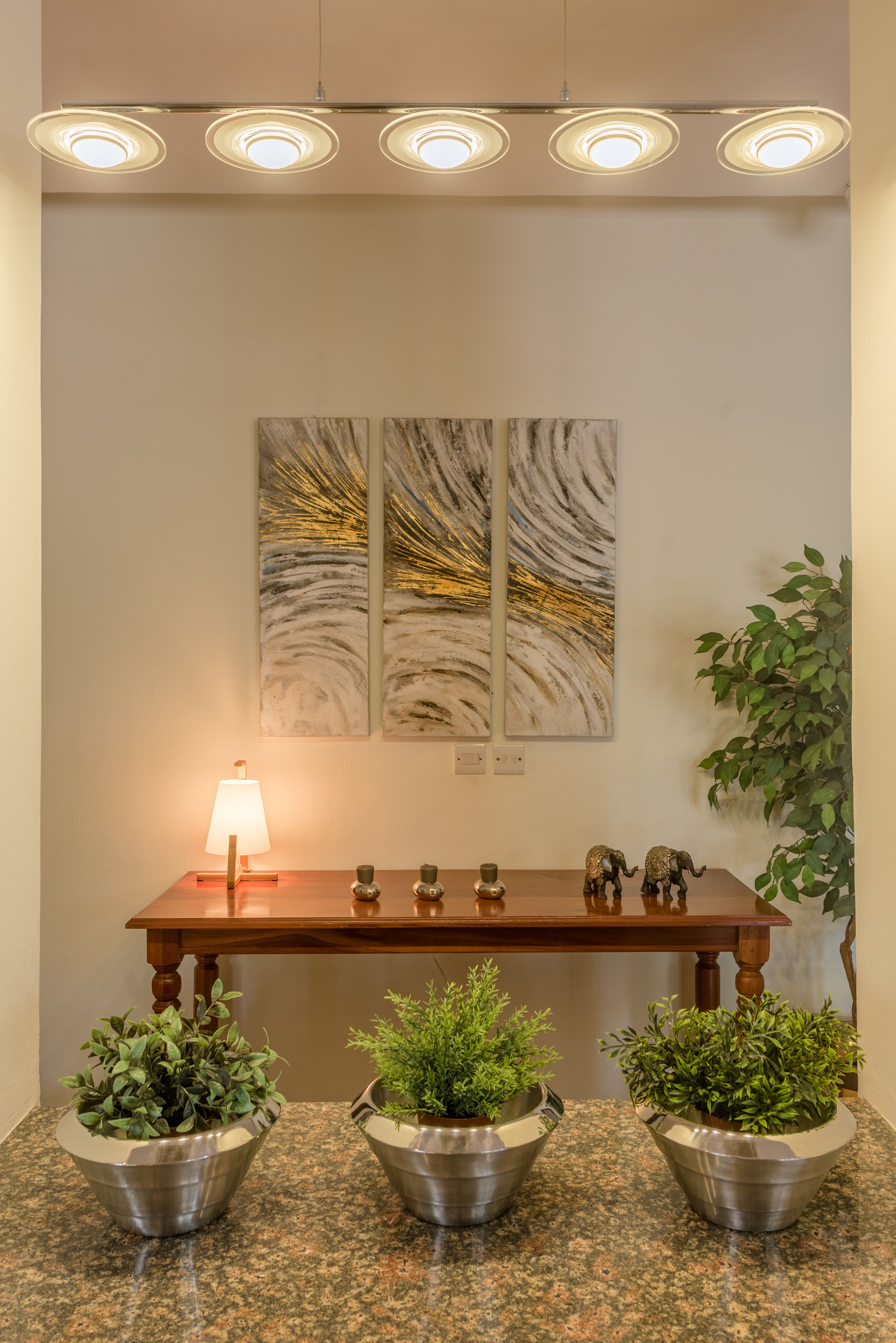 Una habitación bien iluminada con una mesa de madera, tres plantas en macetas, una lámpara y dos cuadros colgados en la pared.