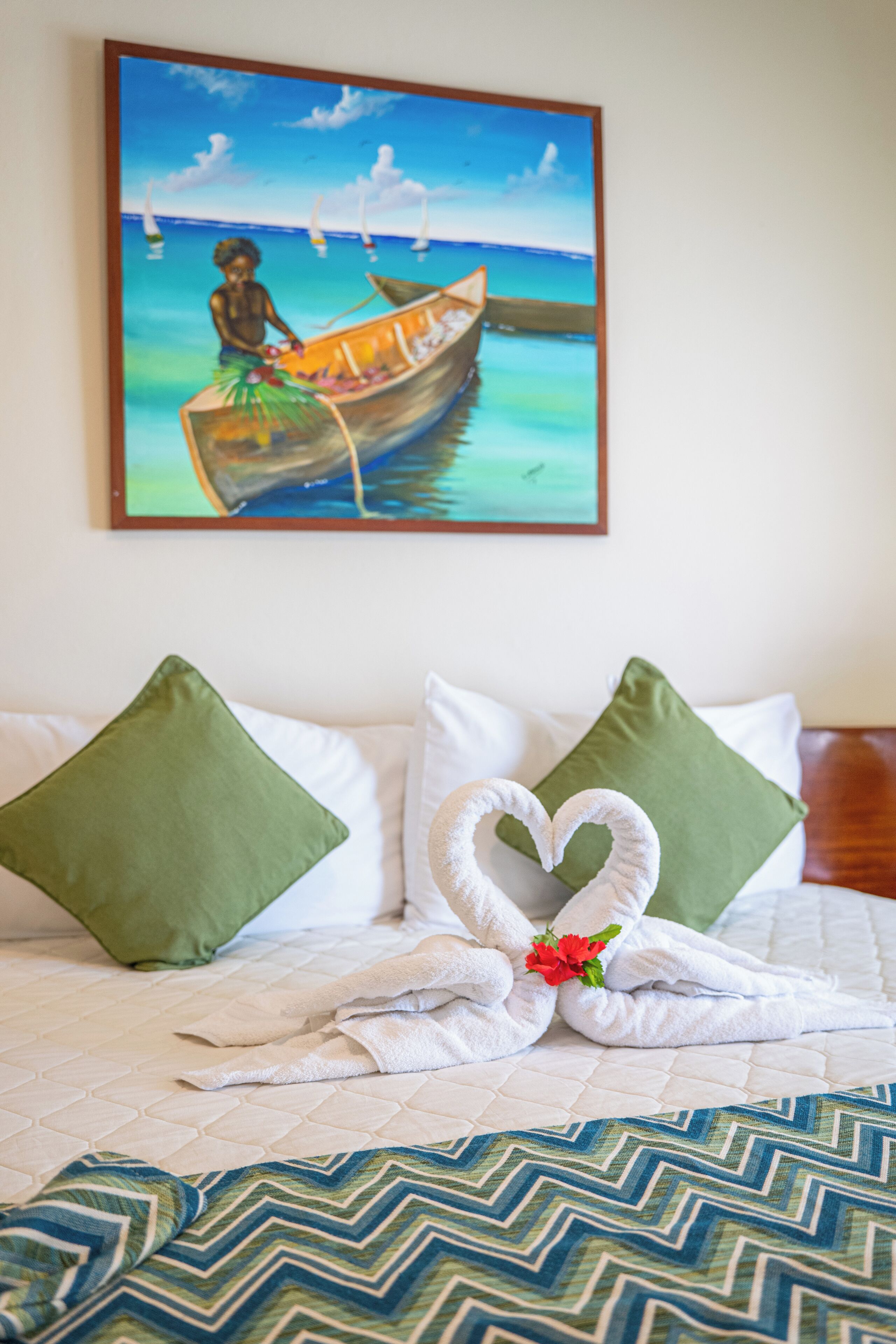 A hotel room with a bed, green pillows, a heart-shaped towel, and a painting of a boat on the wall.