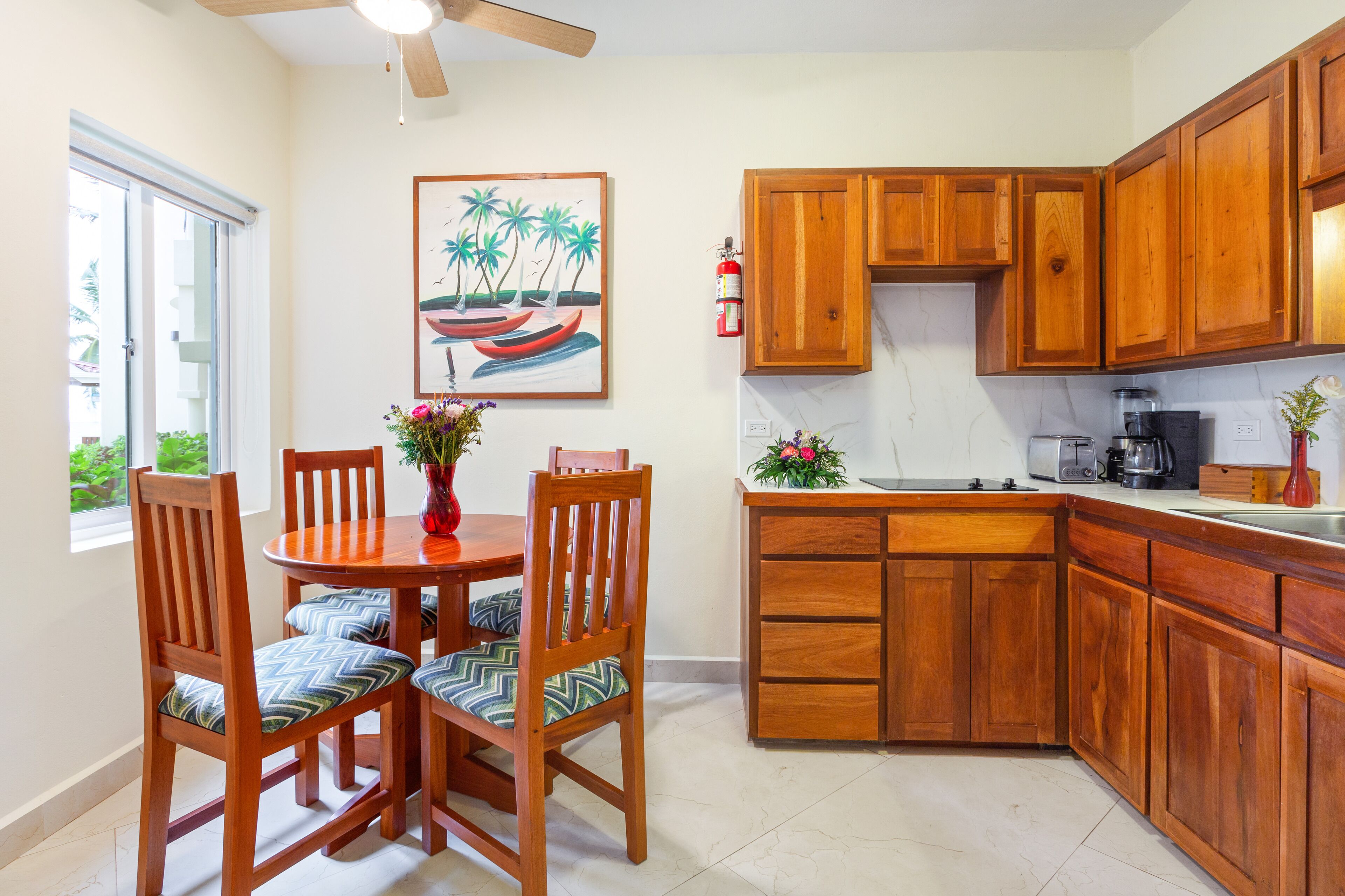 Una cocina con armarios de madera, una mesa de comedor con sillas, un ventilador de techo y una ventana con vistas a la vegetación.