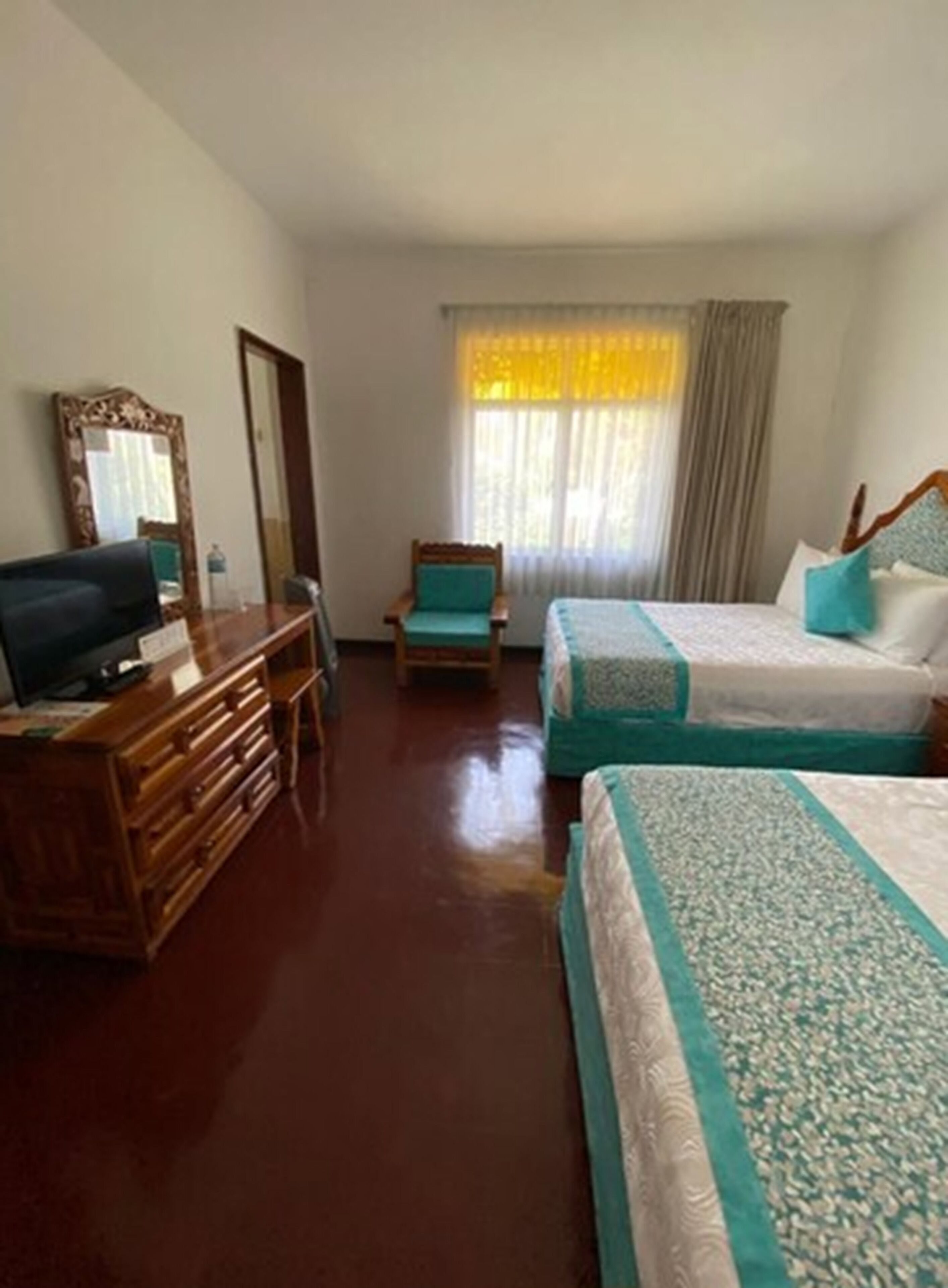 A hotel room with a wooden dresser, a television, a chair, two beds with teal bedding, and a window with curtains.