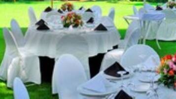 Evento formal al aire libre con mesas redondas cubiertas con manteles blancos, cada una con sillas blancas y centros de mesa.