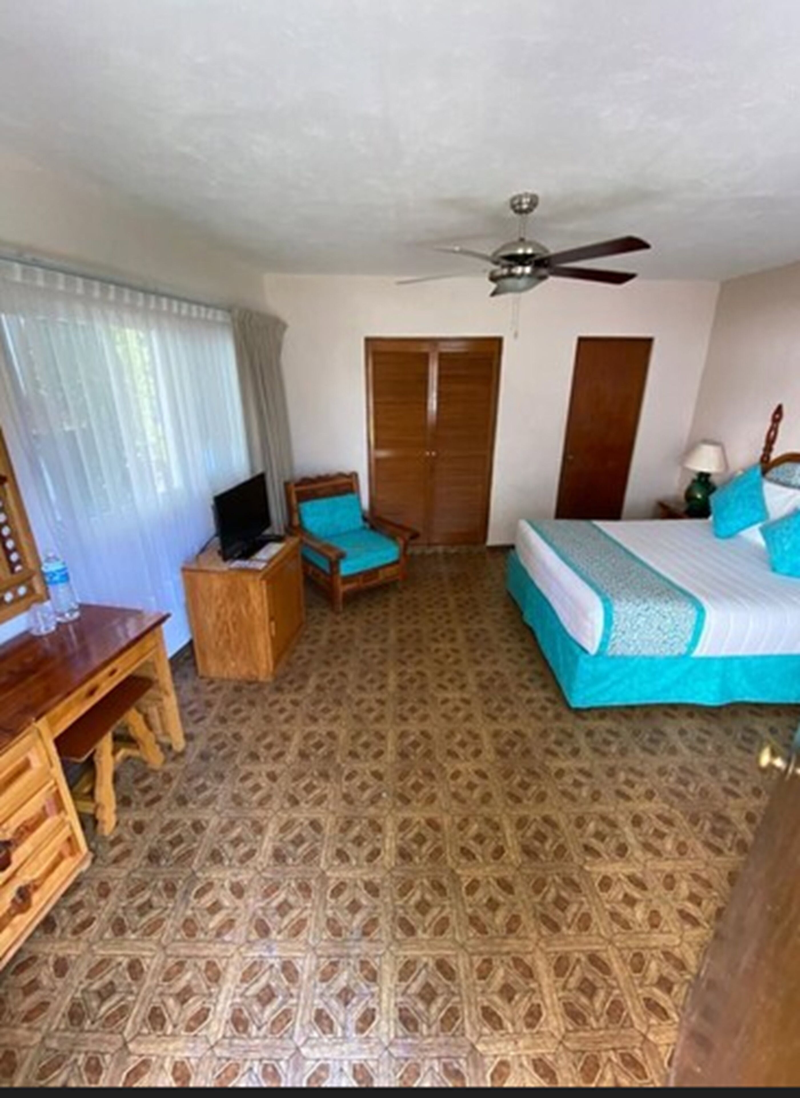 A hotel room with a patterned floor, a bed with blue bedding, a desk with a chair, a television, and a wardrobe.