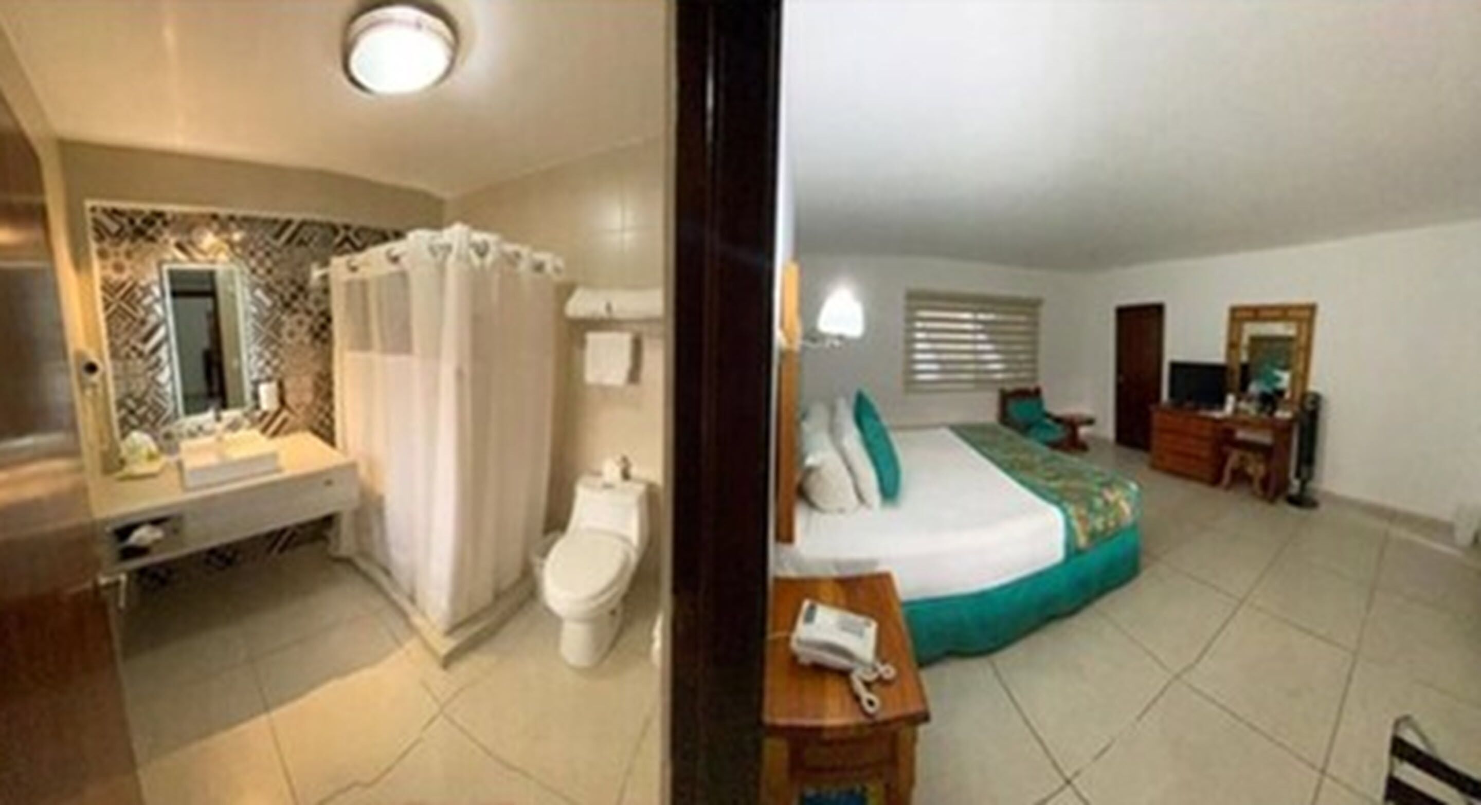 Una habitación de hotel con baño, una cama con cubrecama verde, una mesita de noche y una ventana con persianas.