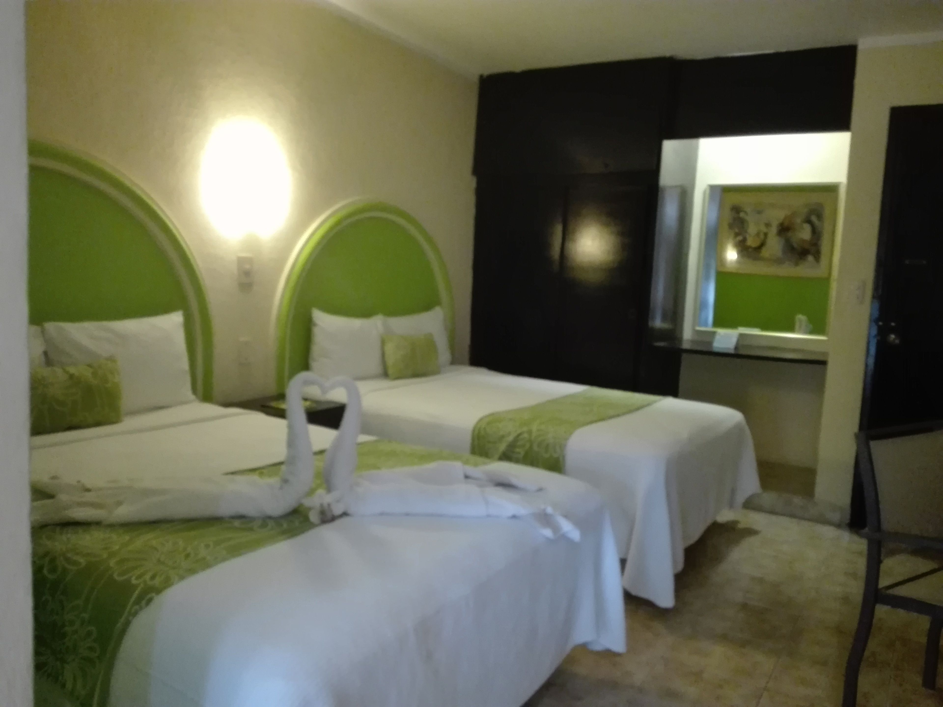 A hotel room with two beds, each with a green headboard and white bedding. There is a mirror on the wall and a chair in the corner.