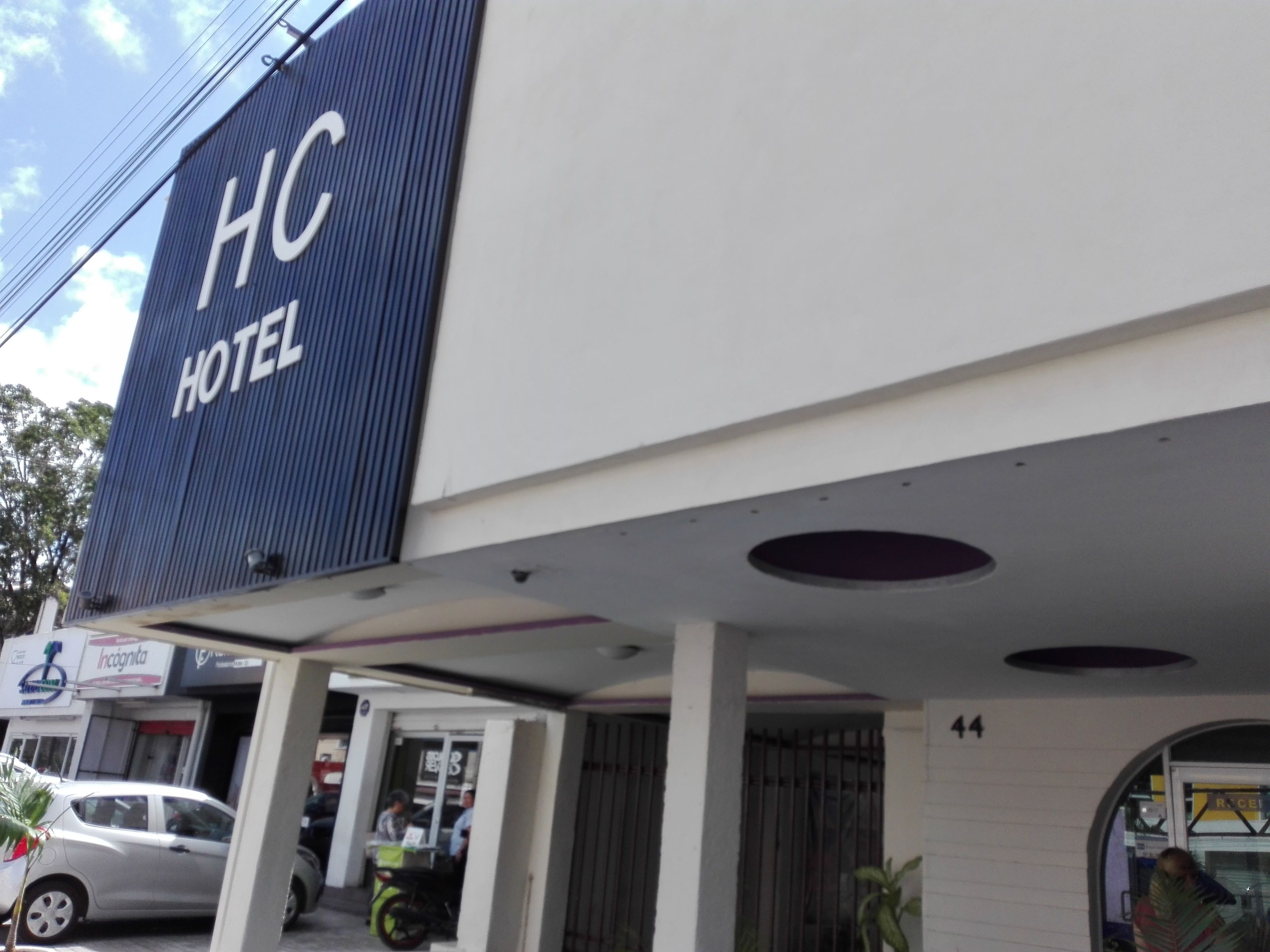 Señal de hotel con la inscripción "HC HOTEL" y la entrada de un edificio con zona cubierta y un automóvil estacionado afuera.