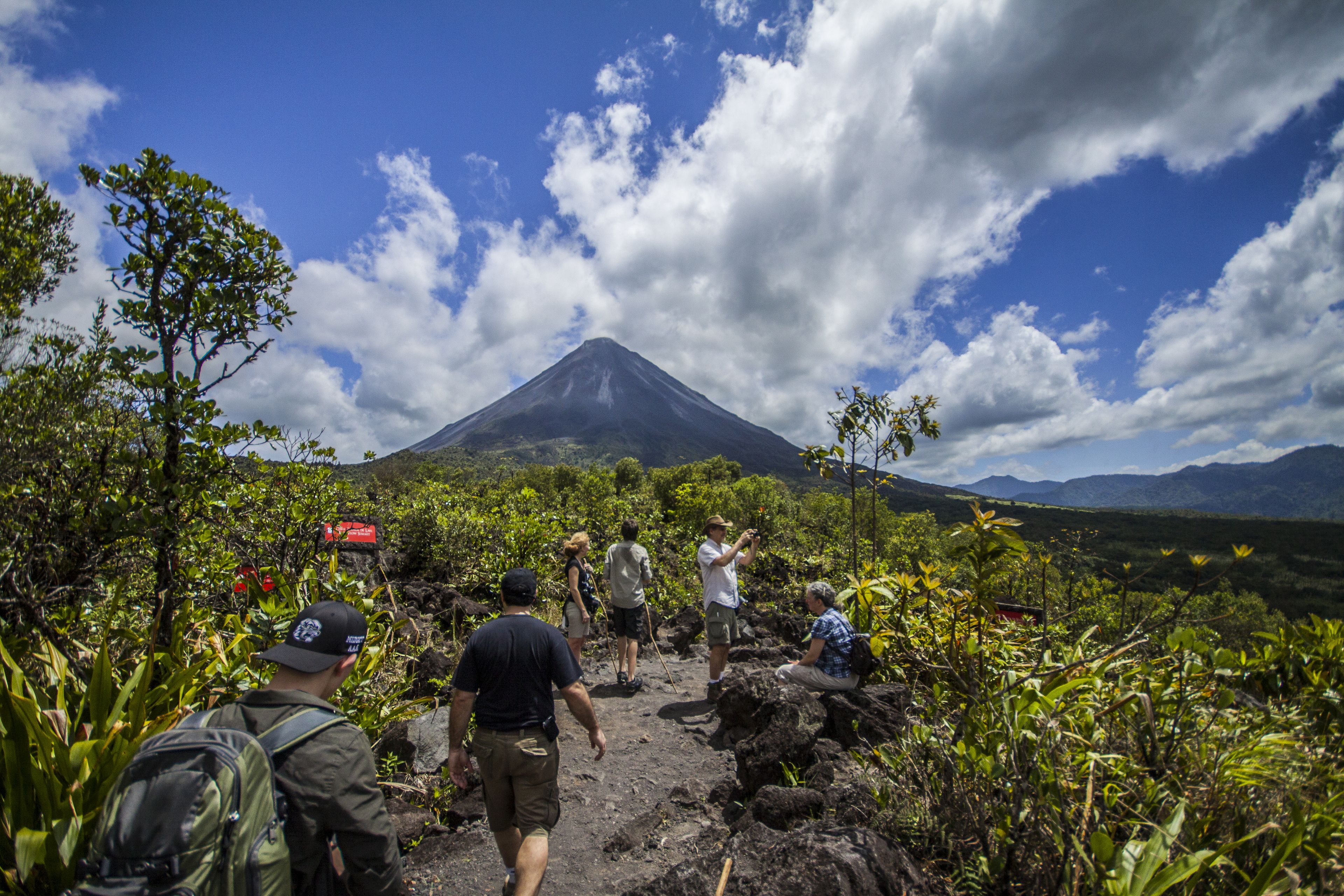 Un groupe de personnes fait de la randonnée sur un sentier rocailleux, avec un volcan en arrière-plan.