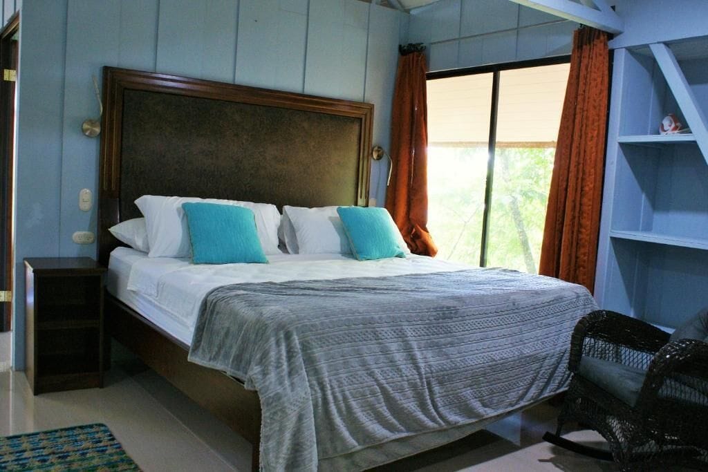 Ein Schlafzimmer mit einem Bett, einem Nachttisch, einem Sessel und einem Fenster mit Vorhängen.