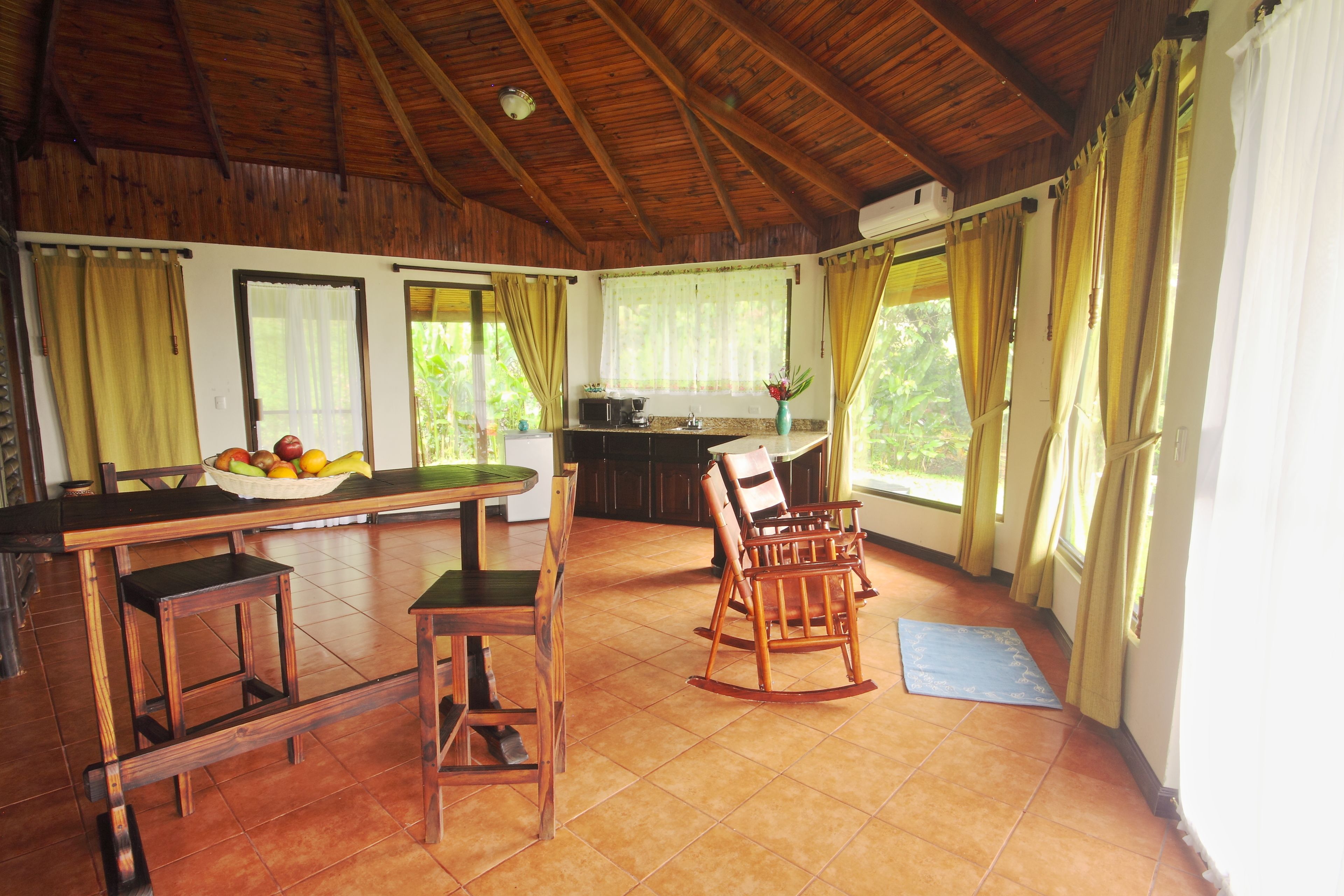 A spacious room with a wooden ceiling, a dining area with a fruit bowl, a rocking chair, and a kitchen area with a window.
