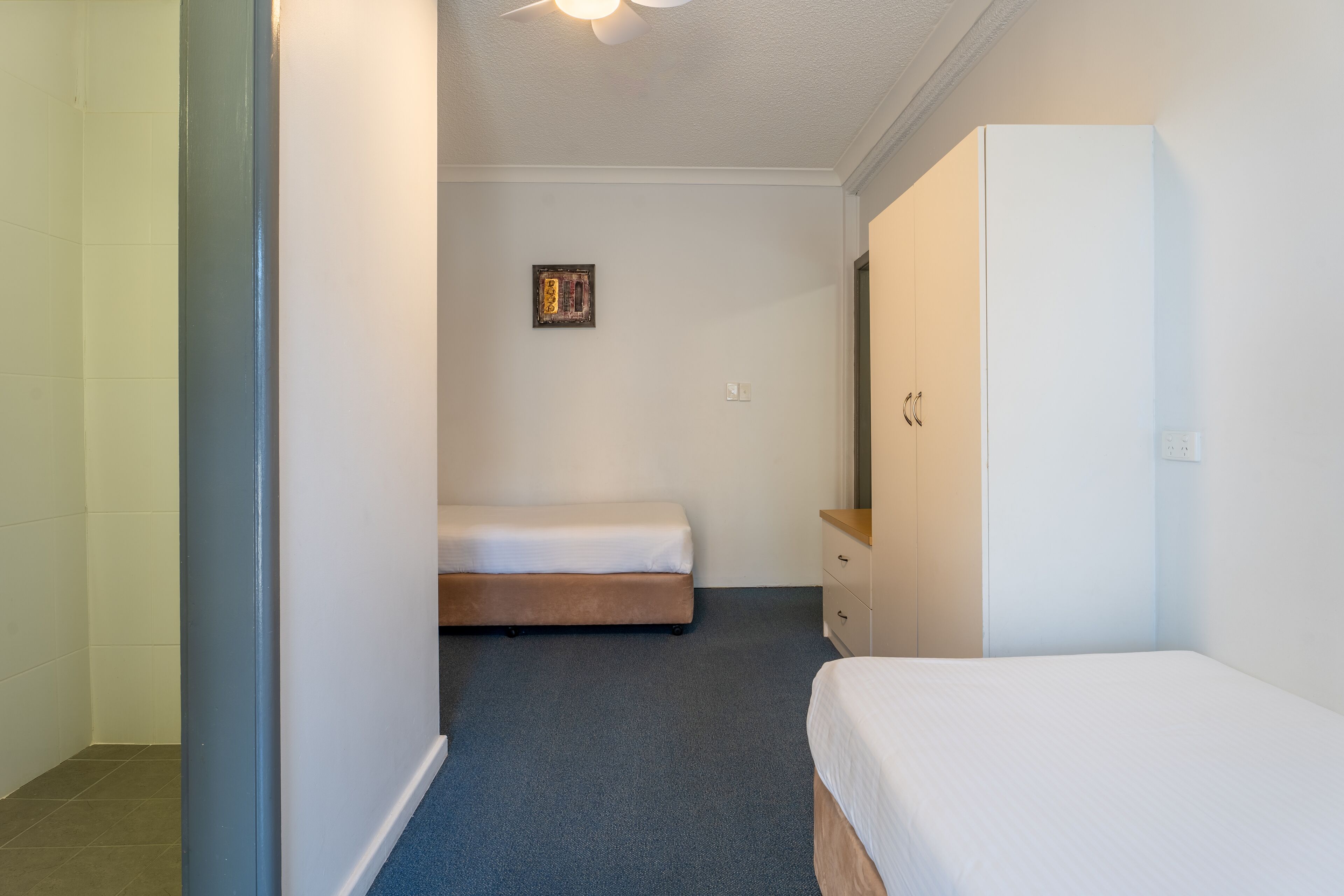 A narrow hallway with a bed on the left, a dresser on the right, and a small framed picture on the wall.