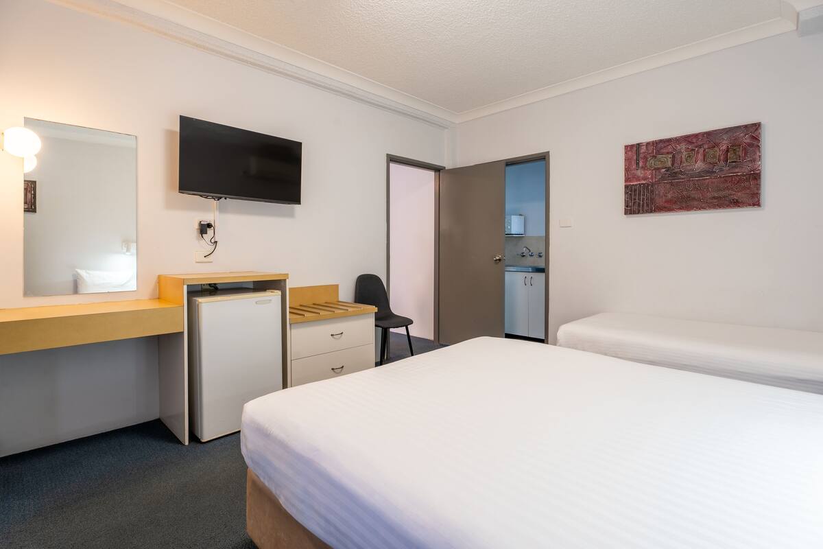A hotel room with a bed, a desk with a computer, a TV, a small fridge, and a door leading to a bathroom.