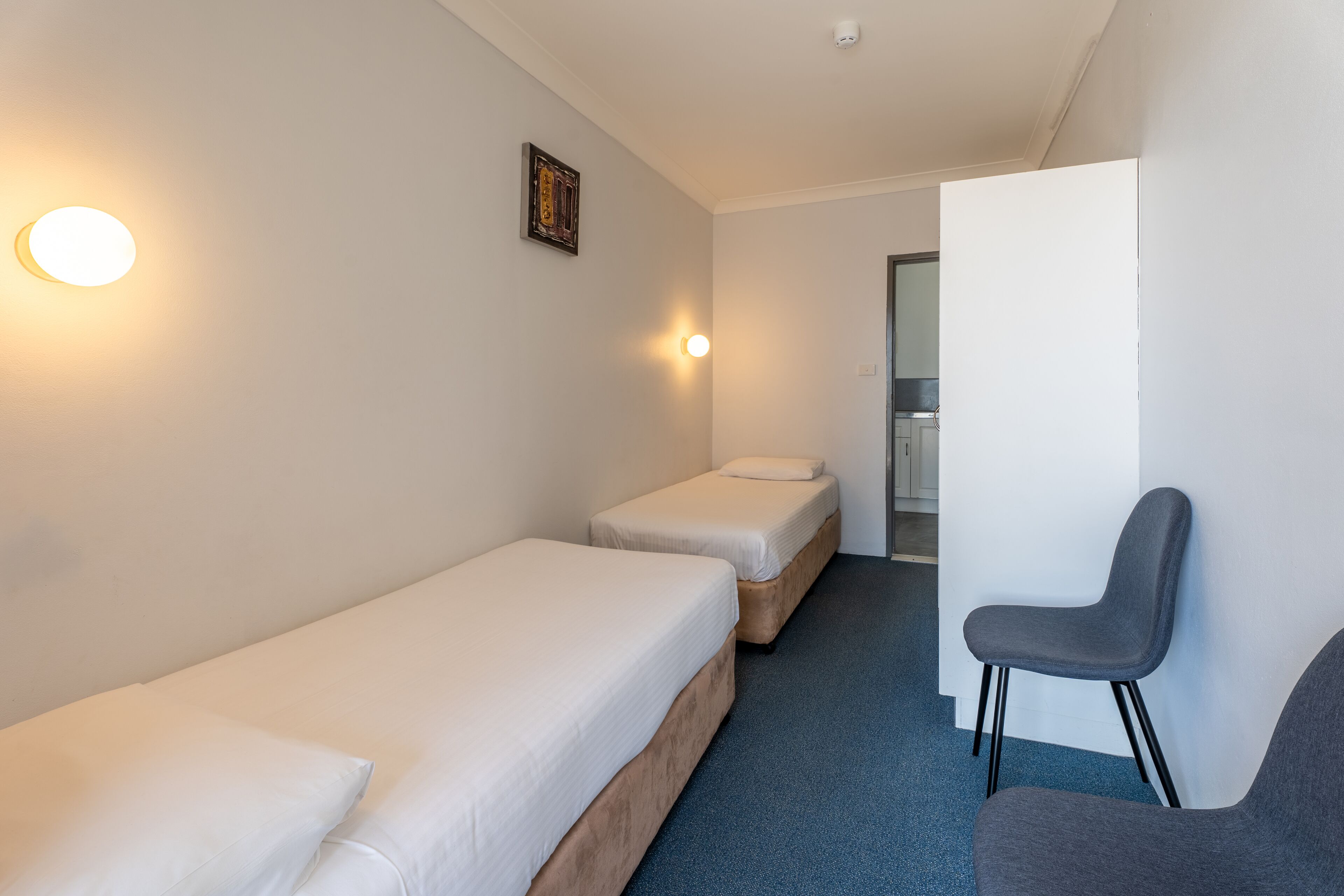 A small, clean room with two single beds, a chair, and a wall-mounted light fixture.